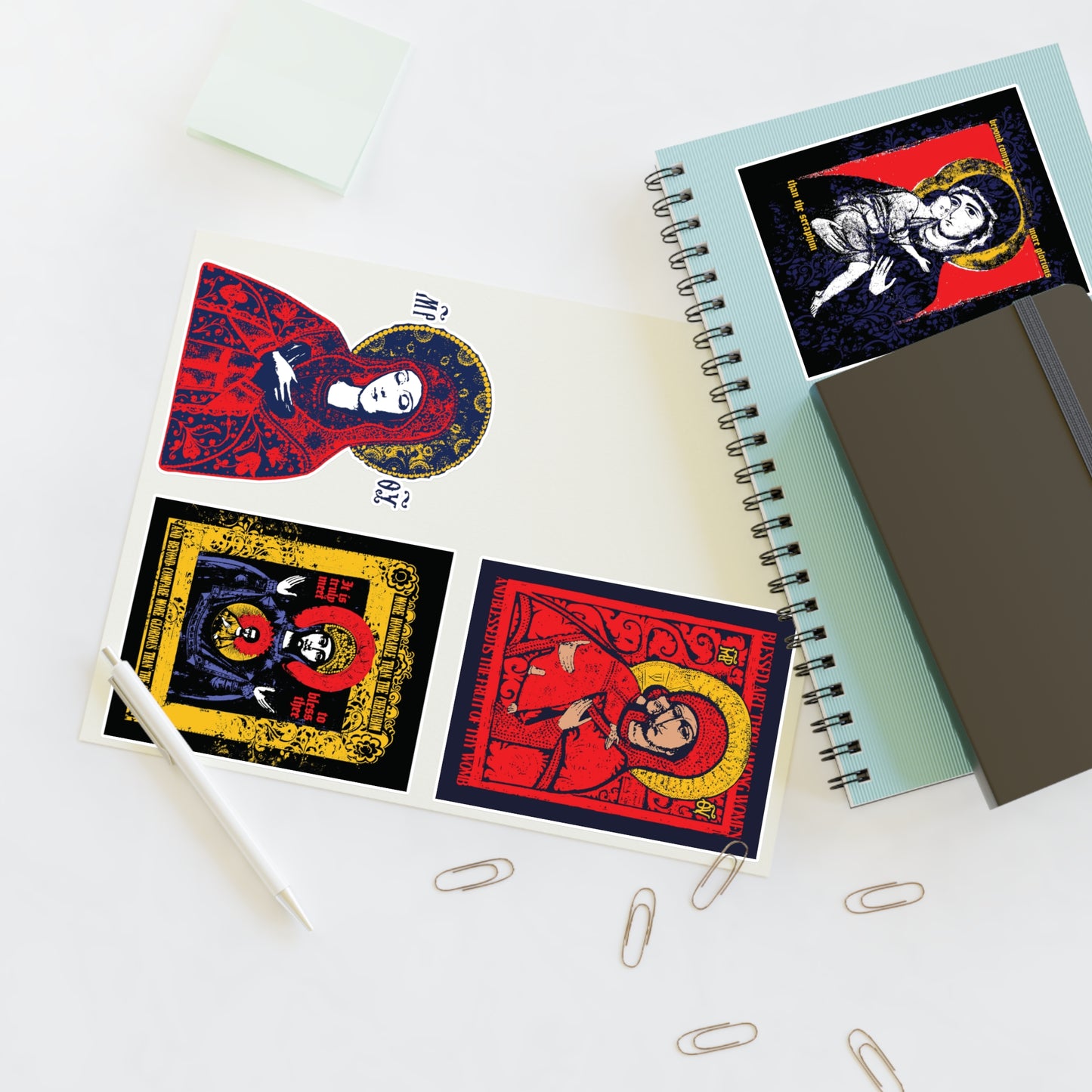 A desk with notebooks, a pen, and paper clips showcases open sketchbooks displaying the Remembrance of Death Theotokos IconoGraphic Sticker Collection No. 1—featuring Vladimir Icon designs, "Joy of All Joys," and "Of the Sign" Orthodox Christian stickers.