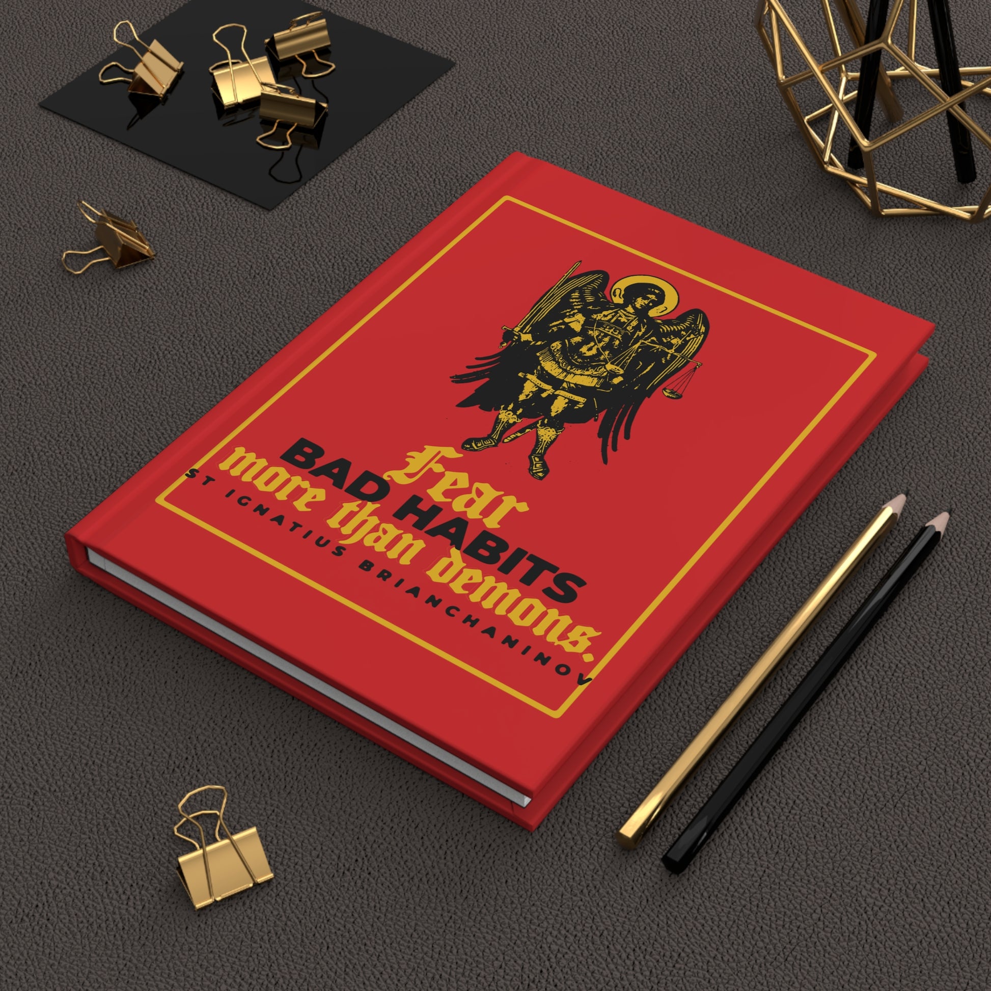 A red "Fear Bad Habits More Than Demons (St Ignatius Brianchaninov) No. 1" Orthodox Christian hardcover journal lies on a desk with pens and gold clips nearby.