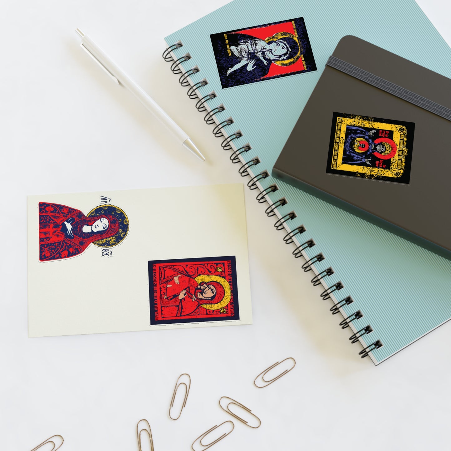 A spiral notebook, a journal, and a postcard with Remembrance of Death Theotokos IconoGraphic Sticker Collection No. 1—Orthodox Christian stickers like the Vladimir Icon—are set on a white surface with a pen and paper clips.