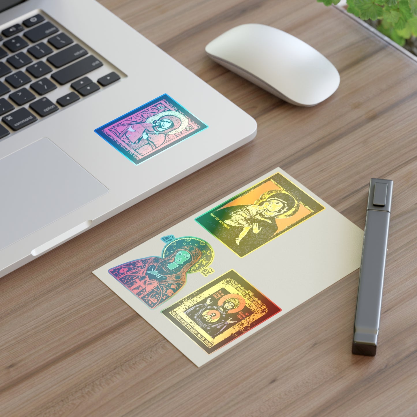 A wooden desk displays a laptop with Remembrance of Death Theotokos IconoGraphic Sticker Collection No. 1, including "Joy of All Joys" and "Of the Sign" Orthodox Christian stickers, plus a white wireless mouse, matching postcard, pen, and a potted plant.