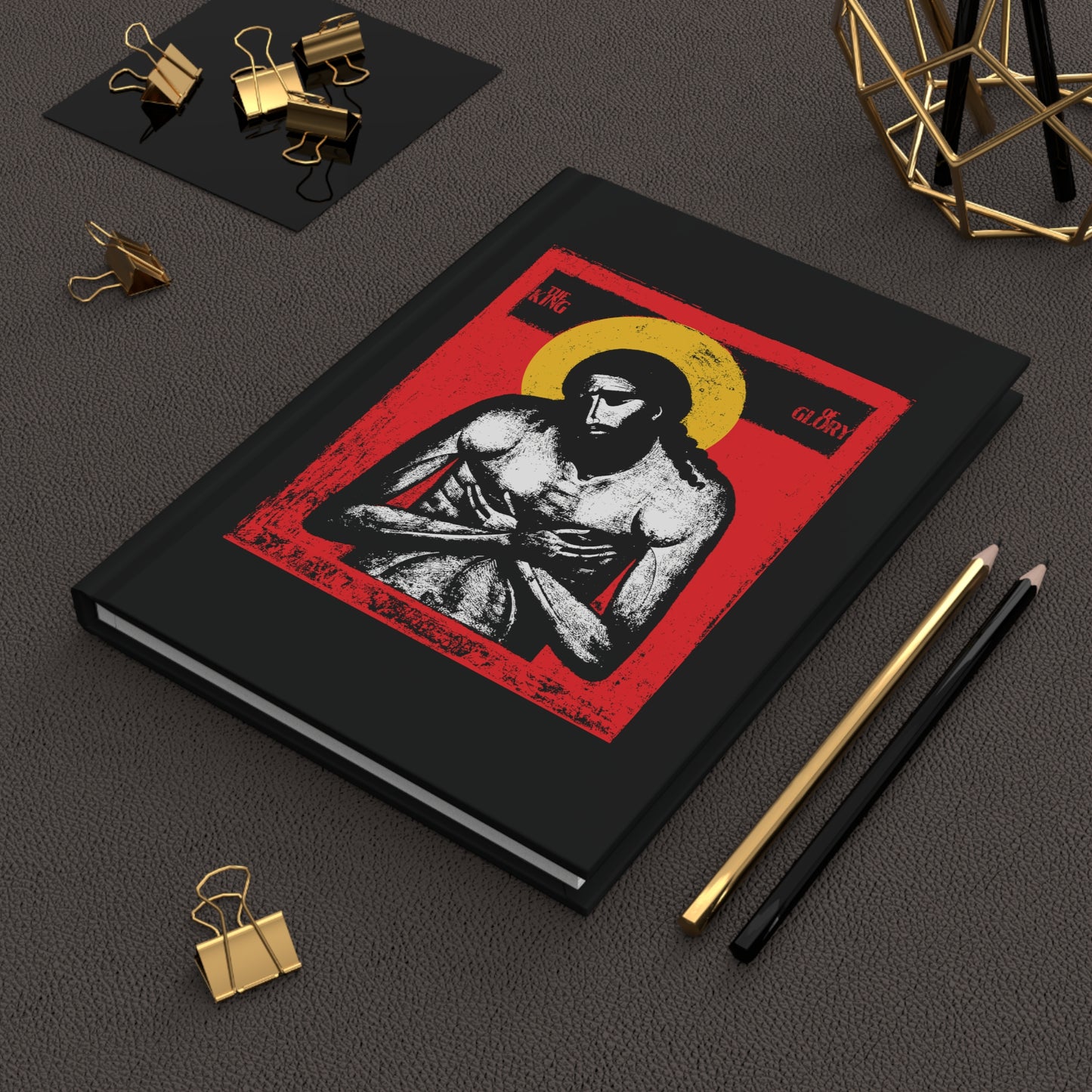 The King of Glory Jesus Christ "Extreme Humility" IconoGraphic Orthodox Hardcover Journal, featuring red, yellow, white, and black, lies beside gold clips, a pencil, and a pen on a dark desk.