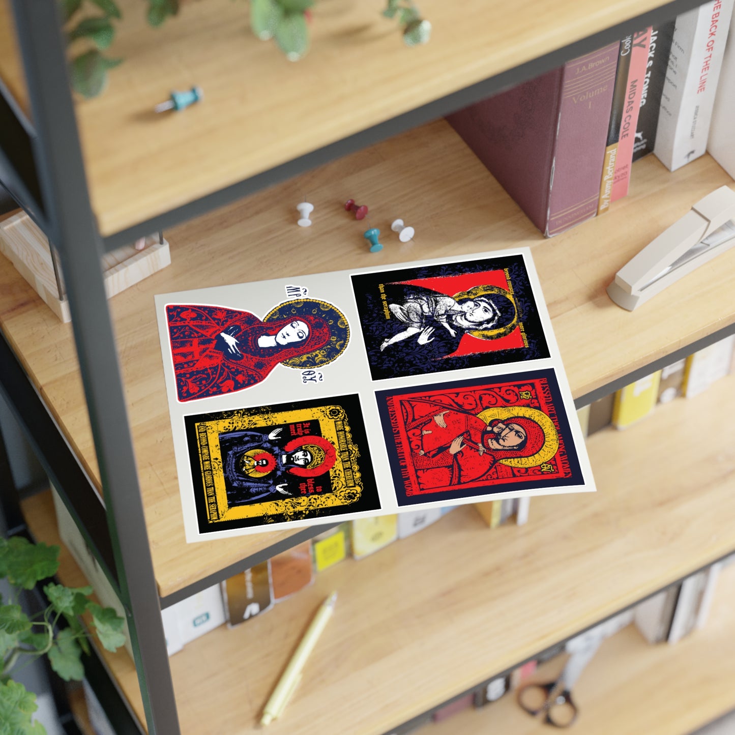 A postcard featuring four vibrant illustrated women’s icons—highlighting the Theotokos IconoGraphic Sticker Collection No. 1 by Remembrance of Death—rests on a wooden shelf beside books, pushpins, a stapler, and green plants.