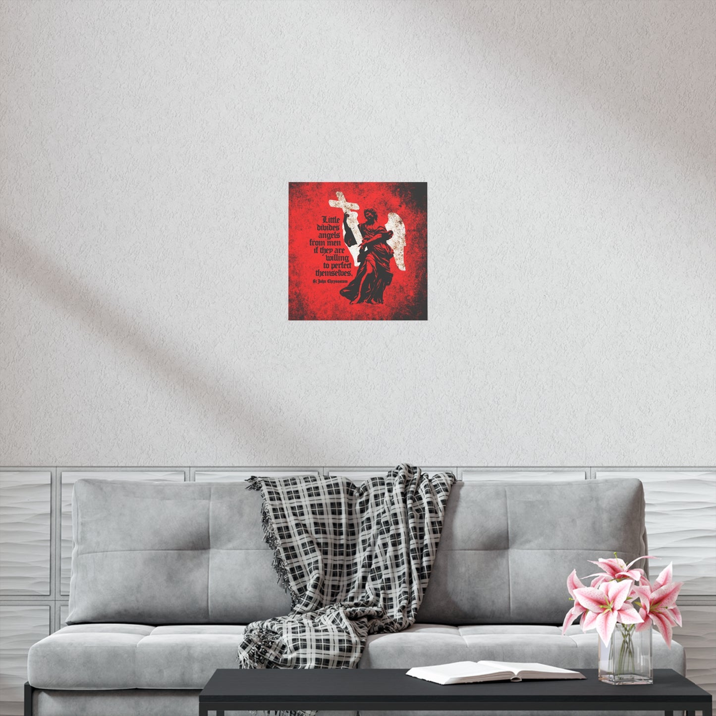 A gray couch with a plaid throw, pink flowers, and an open book sits below the "Little Divides Angels from Men" Orthodox Christian art poster by St John Chrysostom on a white wall, bringing inspiration to the serene space.