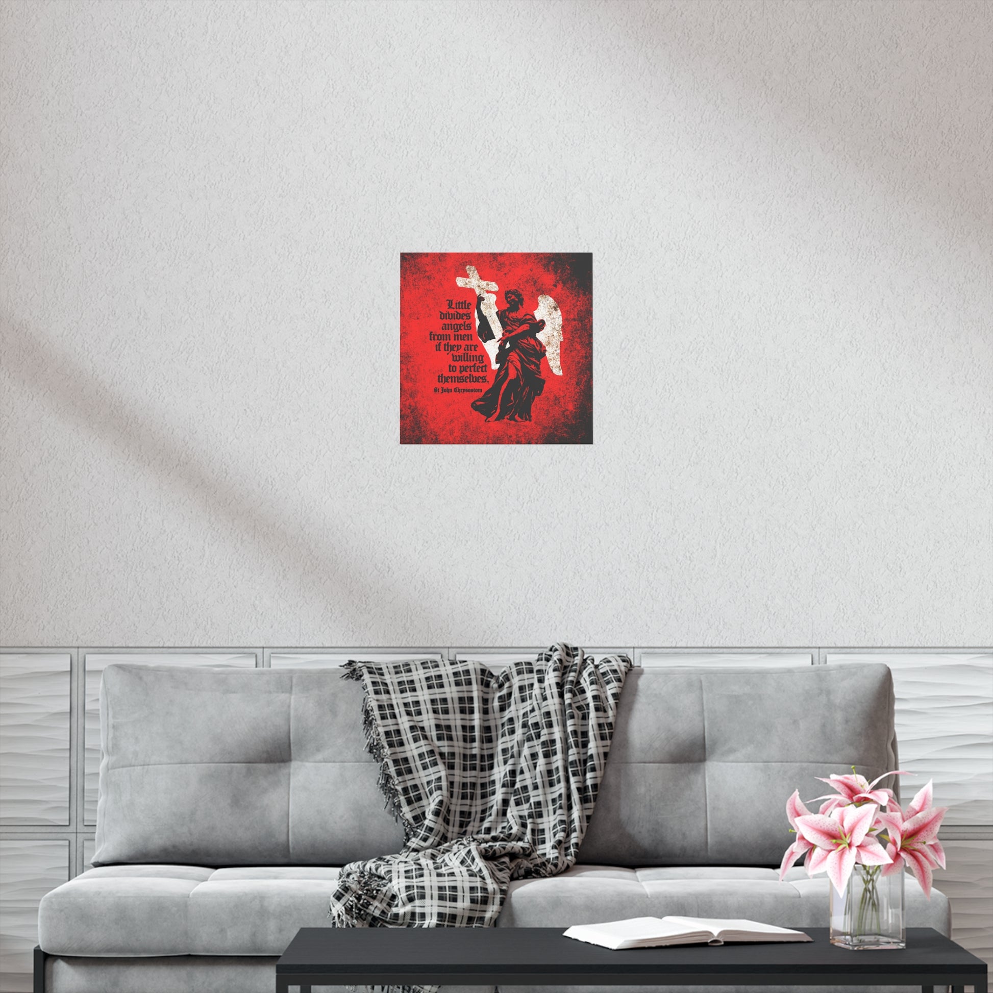 A gray couch with a plaid throw, pink flowers, and an open book sits below the "Little Divides Angels from Men" Orthodox Christian art poster by St John Chrysostom on a white wall, bringing inspiration to the serene space.