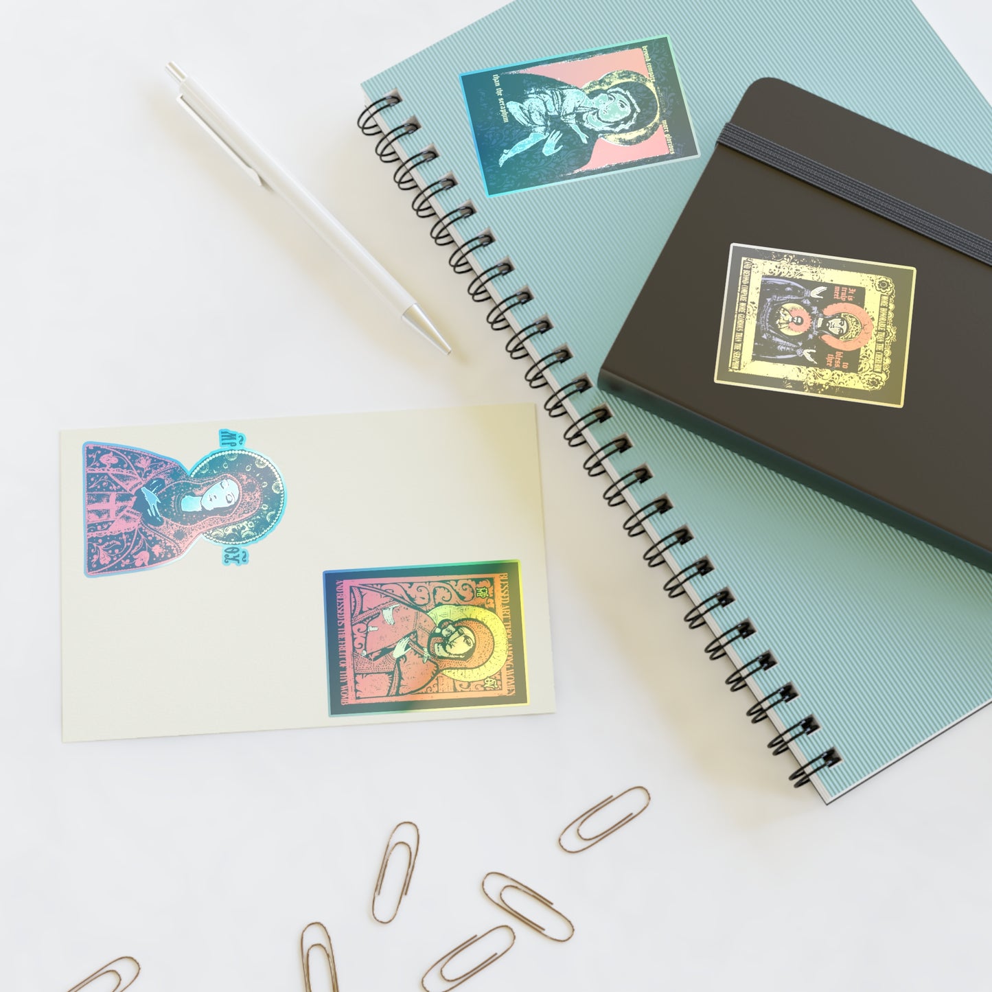 A flat lay shows two spiral notebooks, a card, and a pen on white, all decorated with Remembrance of Death Theotokos IconoGraphic Sticker Collection No. 1—including the Vladimir Icon Design. Several paperclips are scattered nearby.