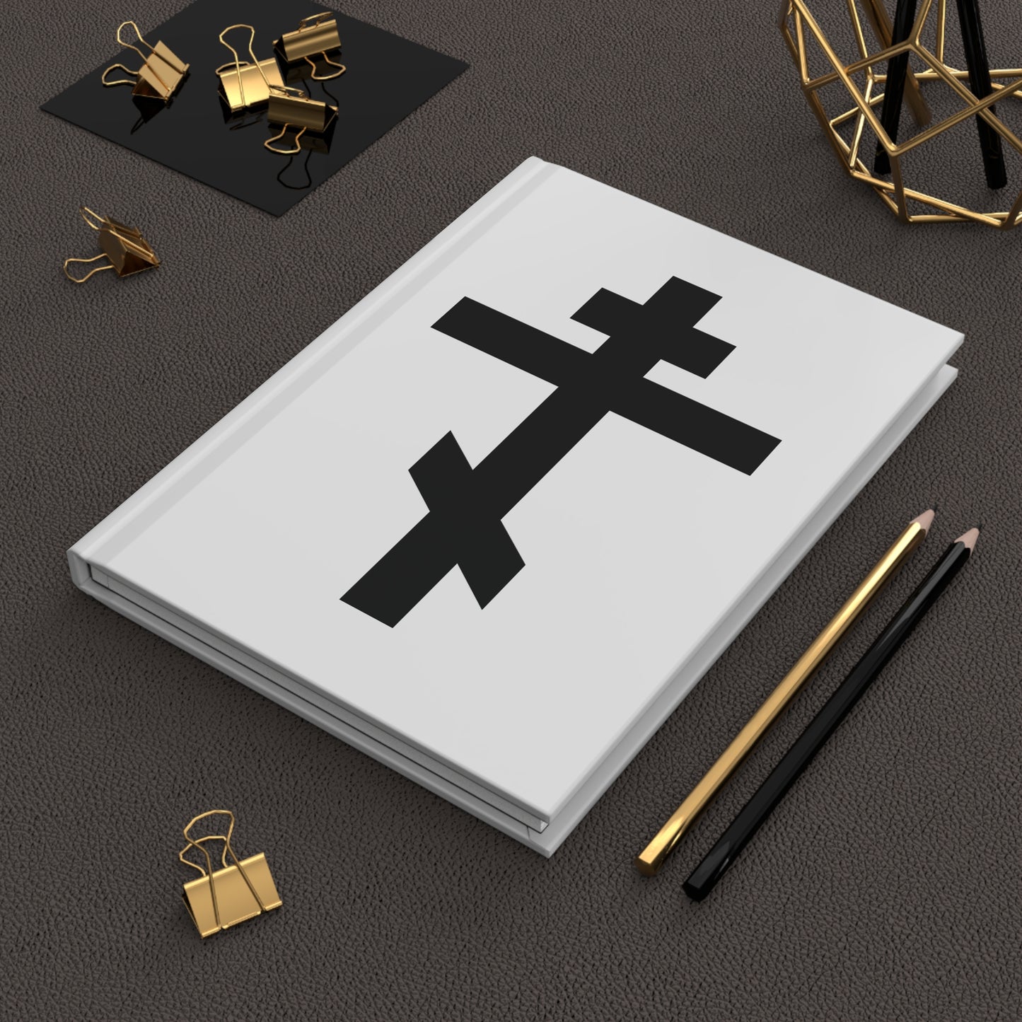 The "Black Orthodox Cross White Background" hardcover journal, featuring Gal 6:14, lies beside gold clips and pencils on a textured desk.