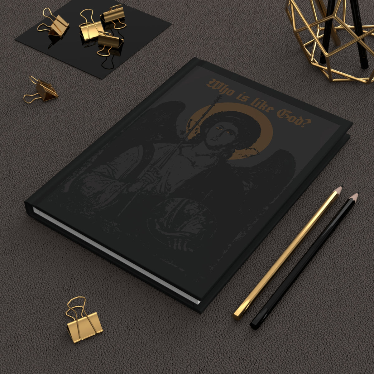 A black & bronze “Who Is Like God? Archangel Michael Icono-Graphic” Goth Christian hardcover journal with a cross on the front left breast sits on a desk next to gold clips, a pen, and a pencil—perfect for any Orthodox Christian.