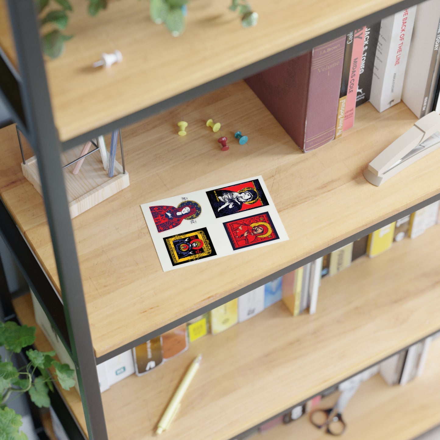 A wooden bookshelf displays books, push pins, a stapler, a small plant, and a sheet of four colorful artistic cards, including a Remembrance of Death Orthodox Christian sticker from the Theotokos IconoGraphic Collection No. 1 (Vladimir Icon Design).