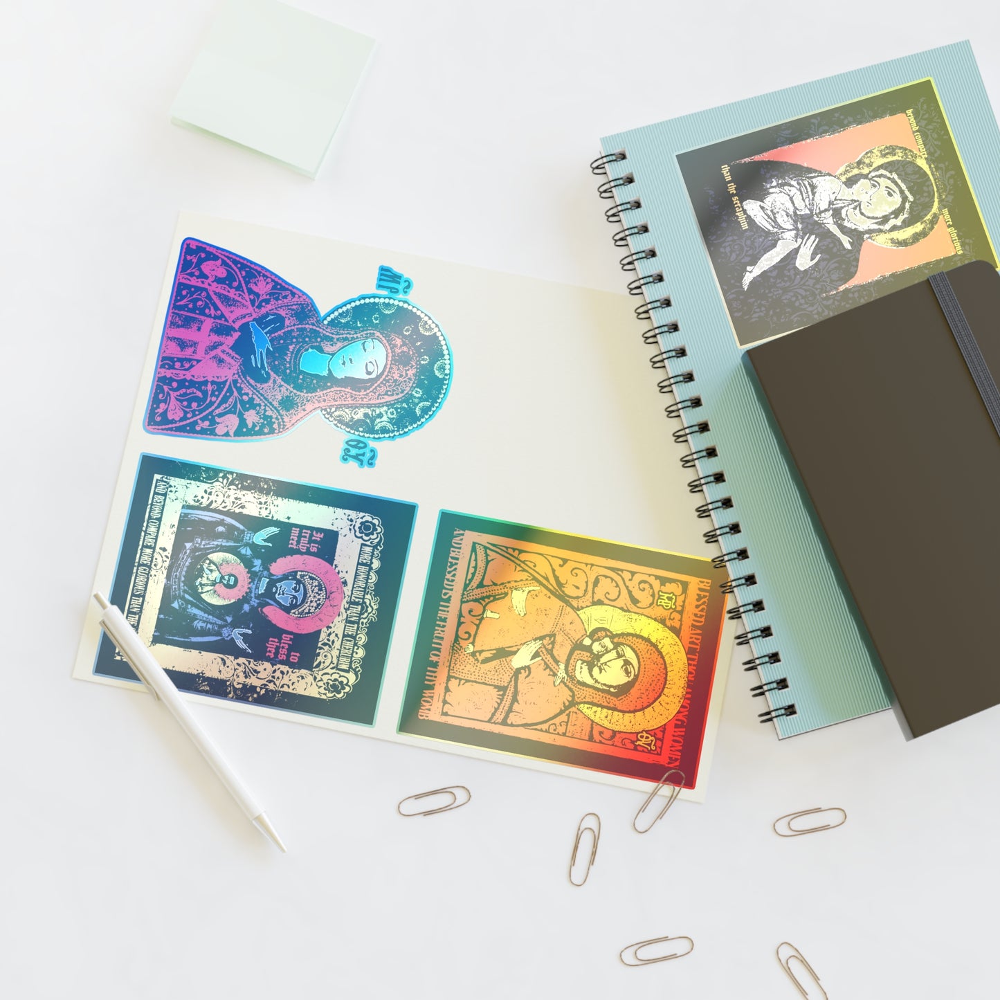 A desk with paper clips, a white pen, sticky notes, blue and black notebooks, and an open book featuring Remembrance of Death Theotokos IconoGraphic Sticker Collection No. 1—Vladimir Icon Design–Orthodox Christian icon stickers.