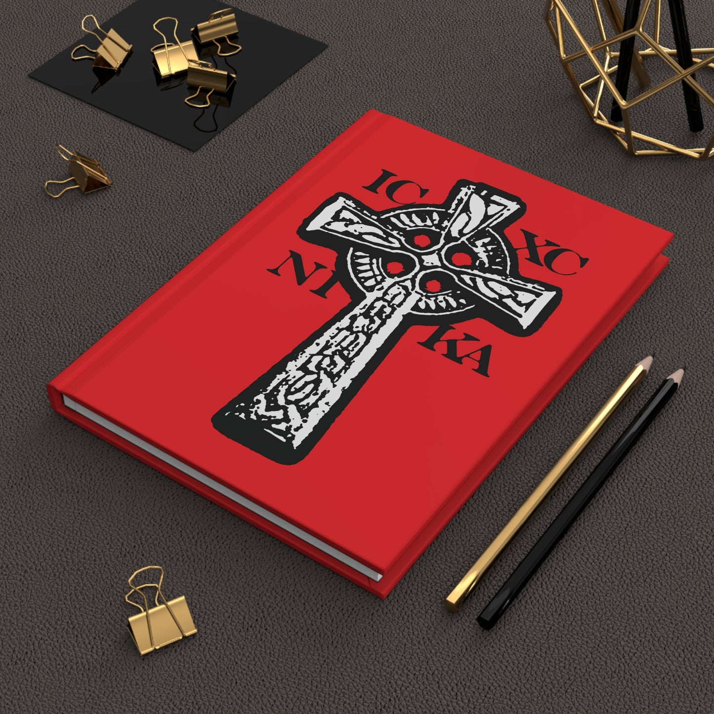 A red ICXC NIKA Celtic Knotwork Cross journal with black and white design, featuring "Jesus Christ Conquers" in Greek, shown beside pencils and binder clips.