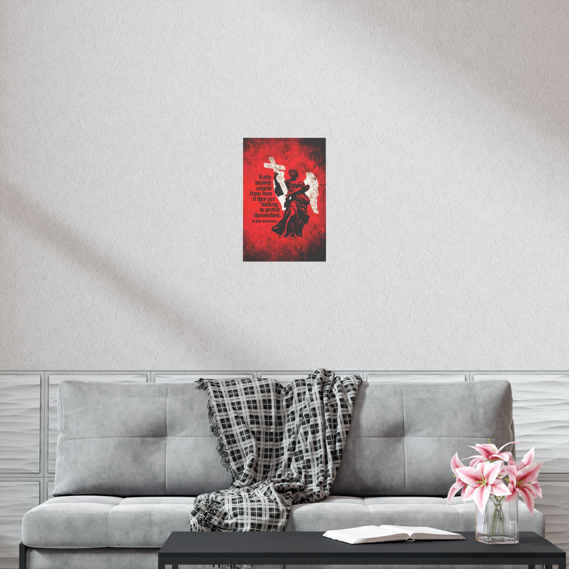A gray couch with a plaid blanket, a book, and pink flowers sits beneath the "Little Divides Angels from Men | Christian Perfection | St John Chrysostom" Orthodox Christian Art Poster, creating a peaceful spot for reflection and grace.