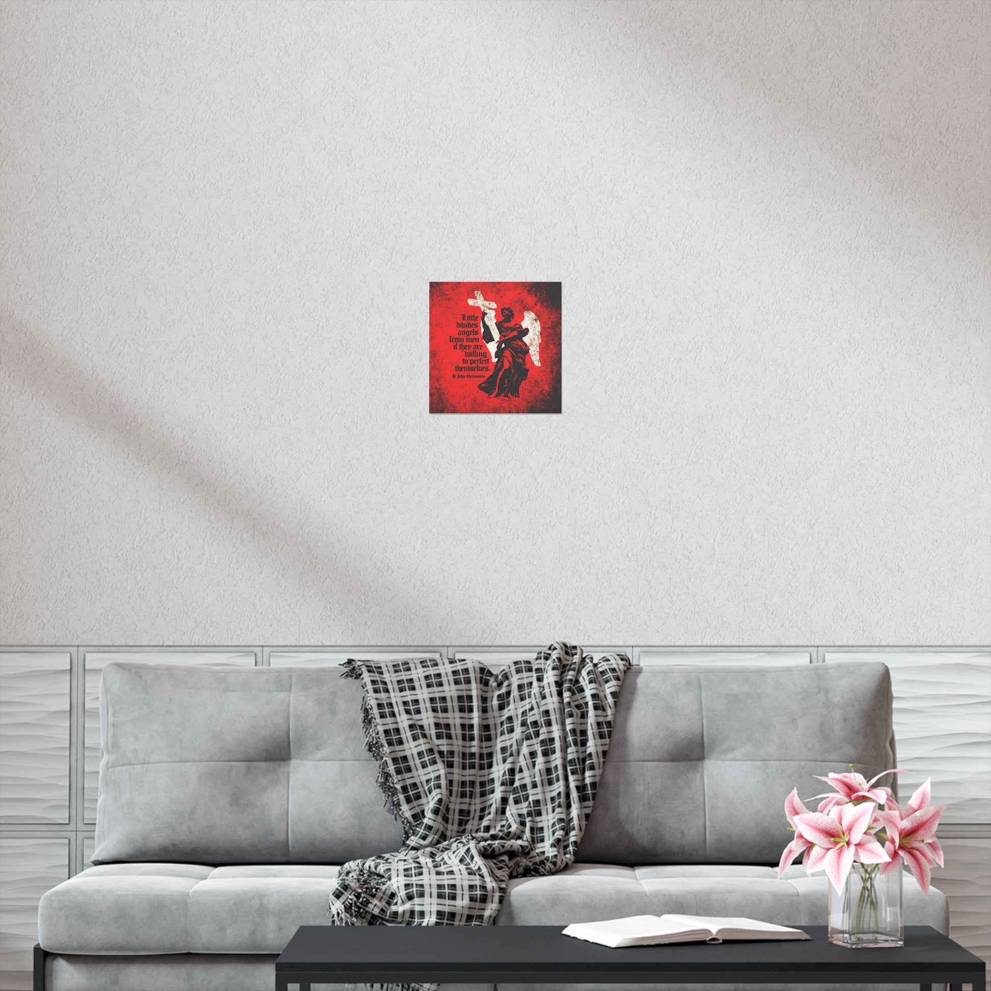 A "Little Divides Angels from Men | Christian Perfection" art poster by St John Chrysostom hangs above a gray sofa adorned with a plaid blanket, flowers, and an open book, inspiring visitors with its Orthodox Christian message.