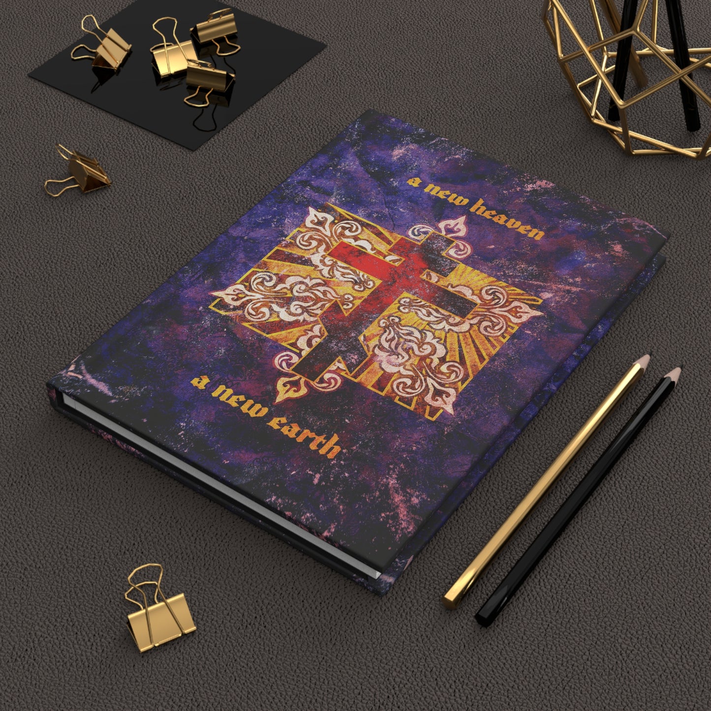 The "A New Heaven/A New Earth" Orthodox Christian Hardcover Journal, featuring a red Orthodox cross on a textured ornate background, sits on a desk surrounded by stationery.