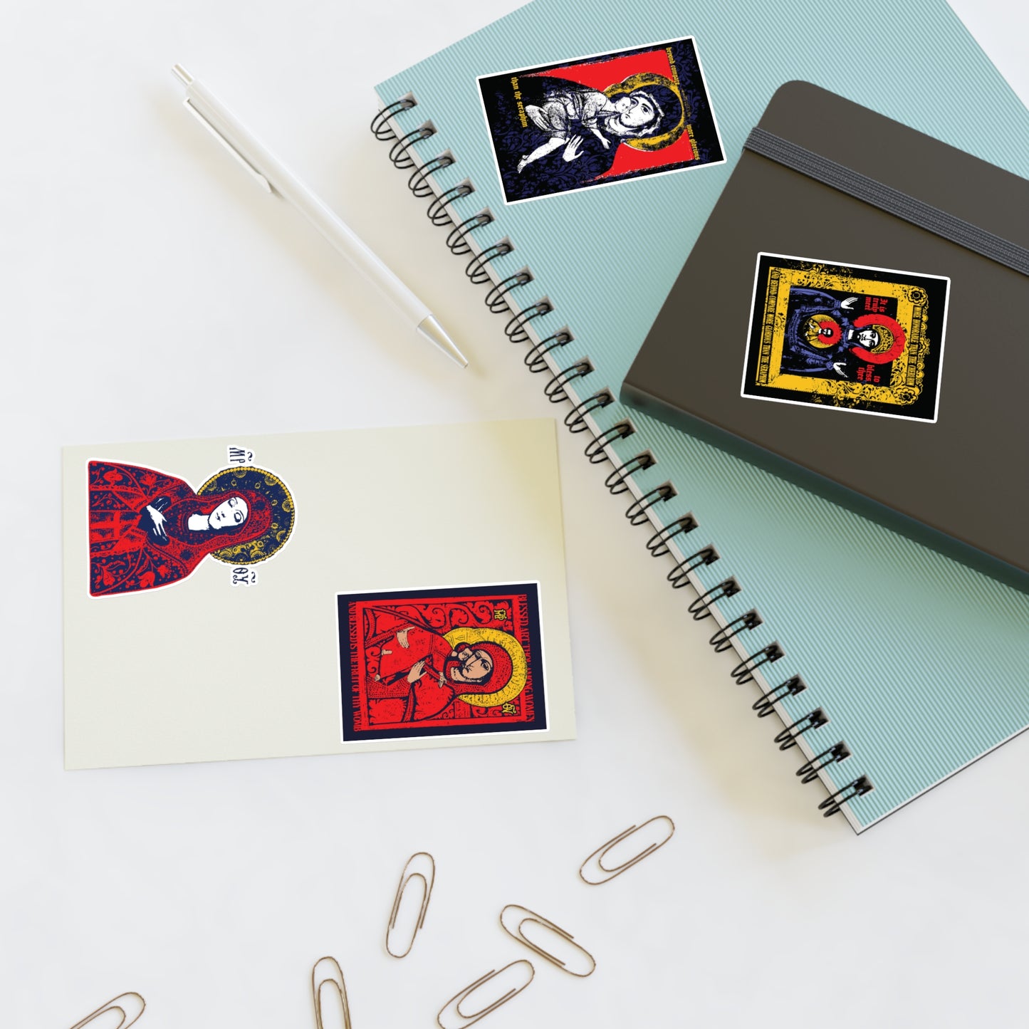 Two spiral notebooks, a pen, and three paper clips sit on a white surface. Each notebook and a postcard feature Remembrance of Death Theotokos IconoGraphic Sticker Collection No. 1 with vibrant Orthodox designs like the Vladimir Icon.
