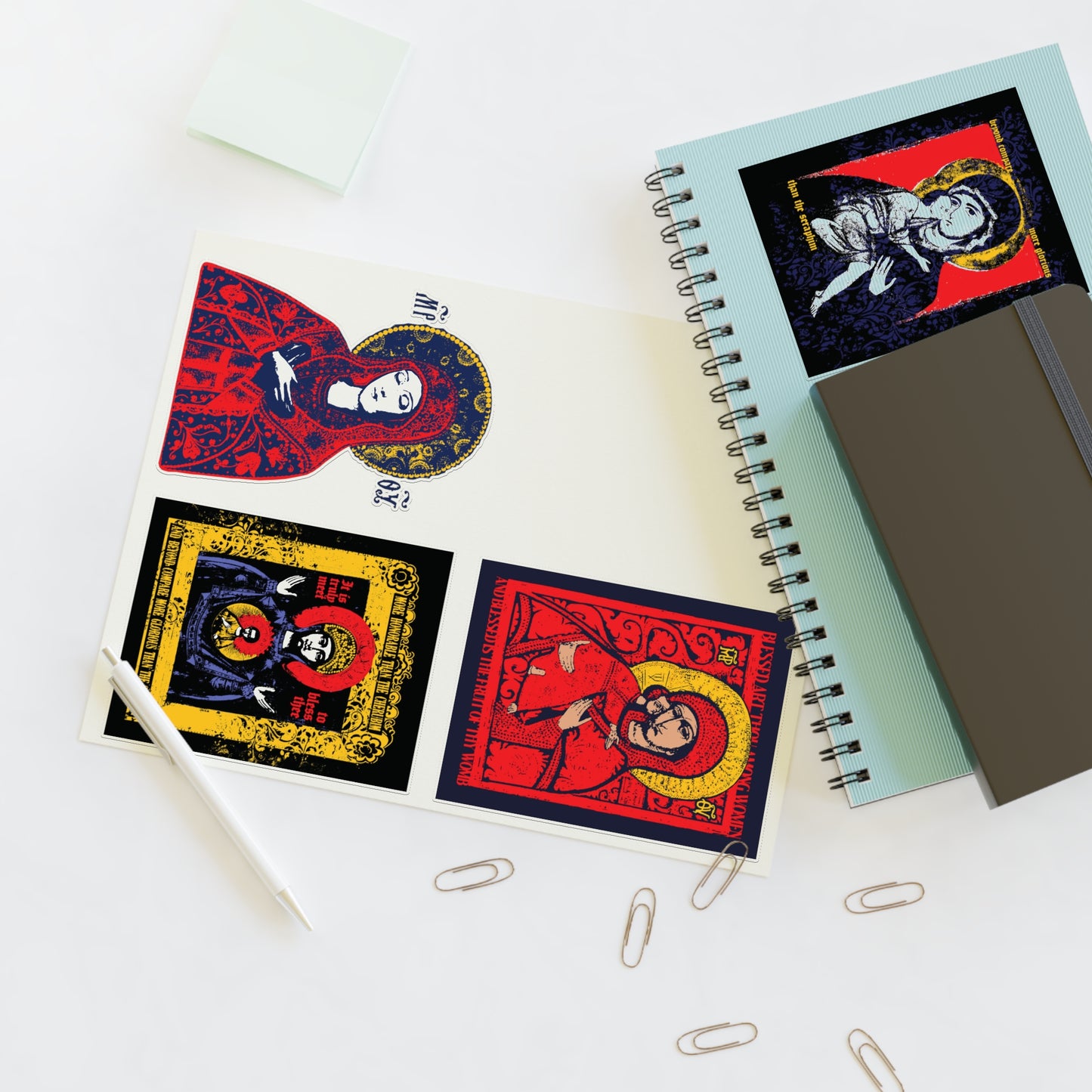 A desk with spiral notebooks, sticky notes, paper clips, a pen, and four Remembrance of Death Theotokos IconoGraphic Sticker Collection No. 1 stickers—each featuring vibrant Orthodox Christian art, including a striking Vladimir Theotokos icon.