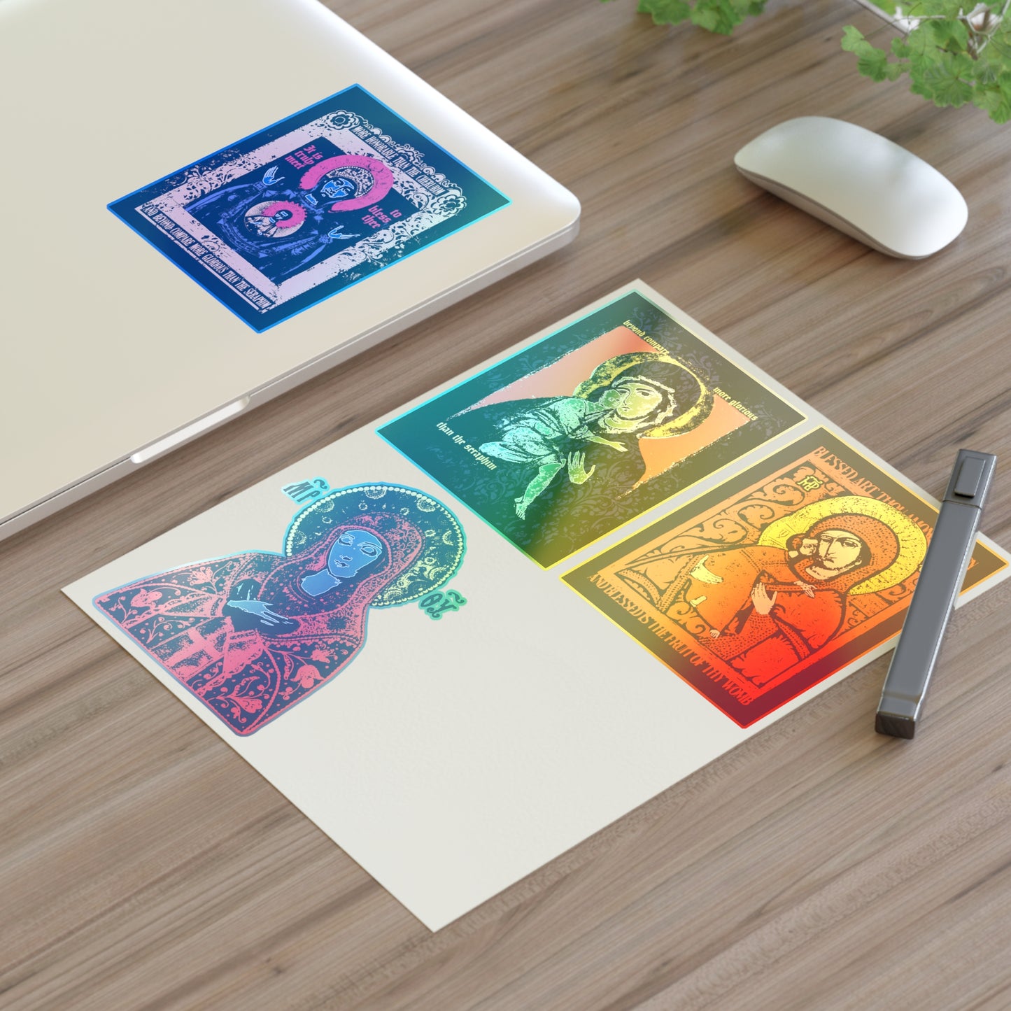 A laptop and mouse rest on a wooden desk with three colorful Remembrance of Death Theotokos IconoGraphic Sticker Collection No.1 Orthodox Christian stickers, including "Joy of All Joys" and "Of the Sign." One sticker is applied to the laptop. A plant appears in the corner.