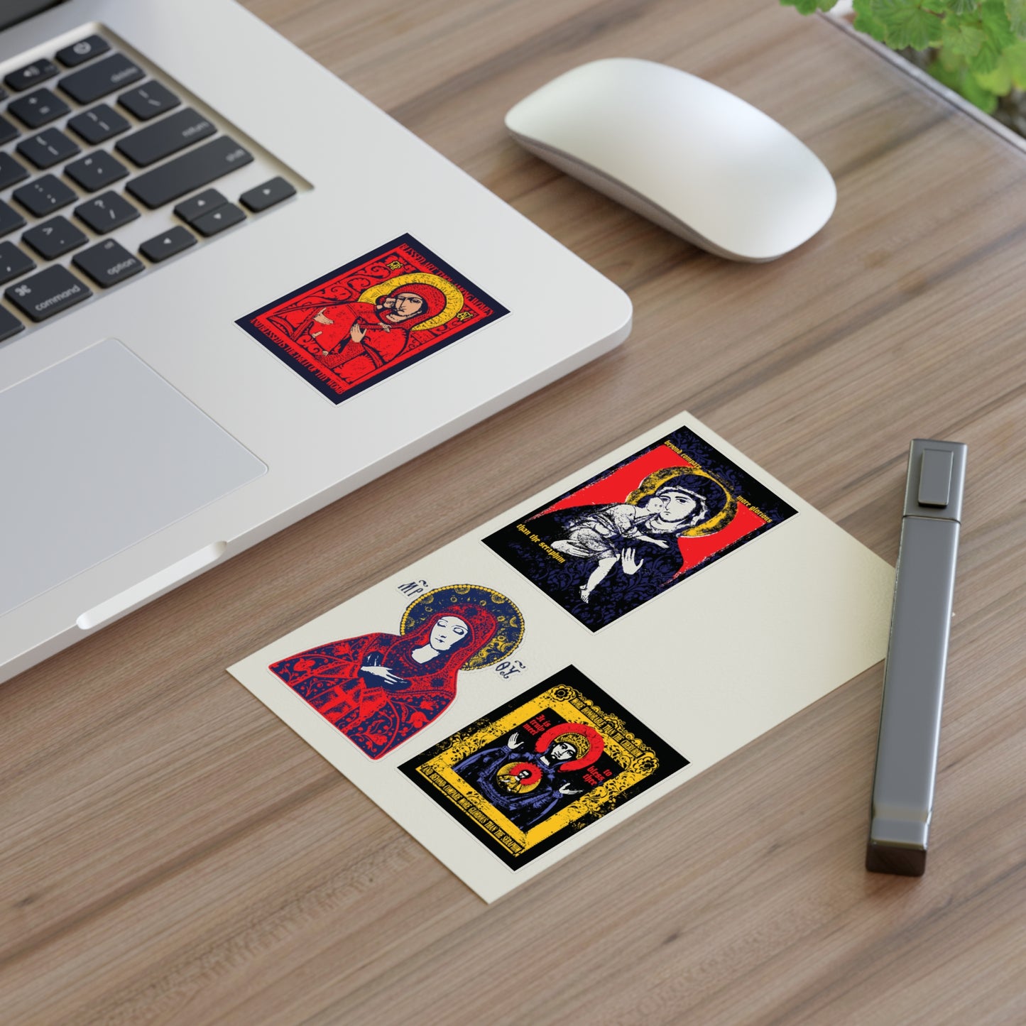 A laptop on a wooden desk with a Remembrance of Death Theotokos IconoGraphic Sticker Collection No. 1, near a postcard of three saint icons, a gray pen, mouse, and a plant partially visible in the corner.