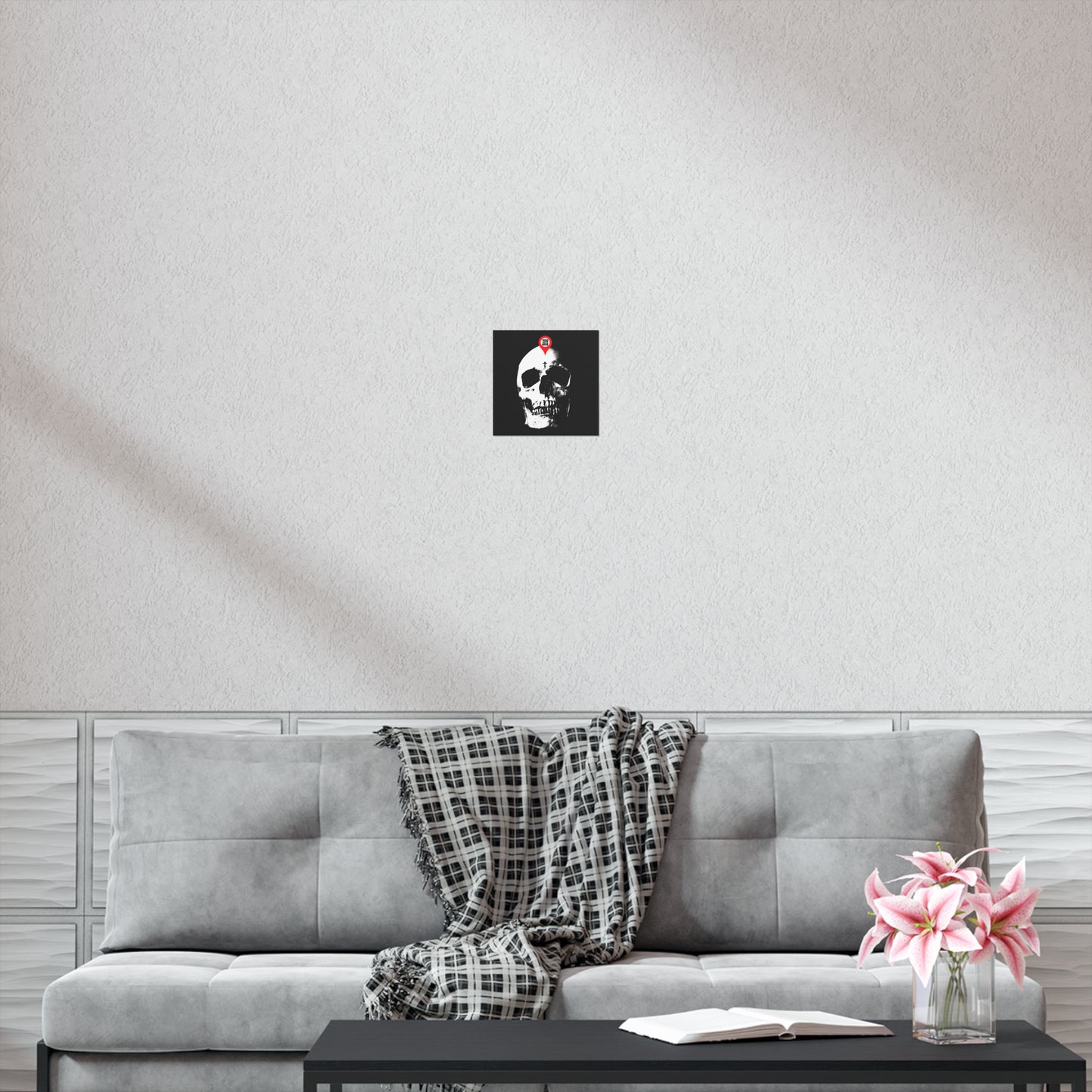The "You Will Be Here Maps Marker on Skull" Orthodox Christian art poster, inspired by memento mori, hangs above a gray sofa with a plaid throw, open book, and pink flowers on the table.