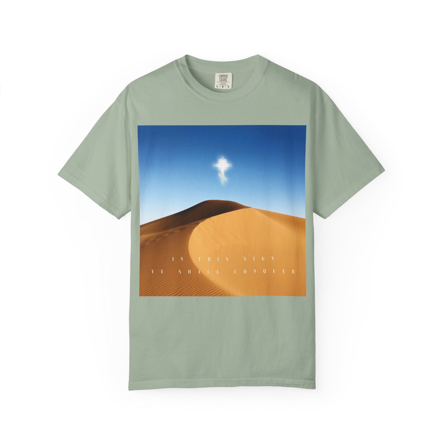 A Bay-colored heavyweight cotton unisex tee featuring a desert sun scene with a glowing cross in the sky symbolizing the Elevation of the Cross, and the text "IN THIS SIGN YE SHALL CONQUER.