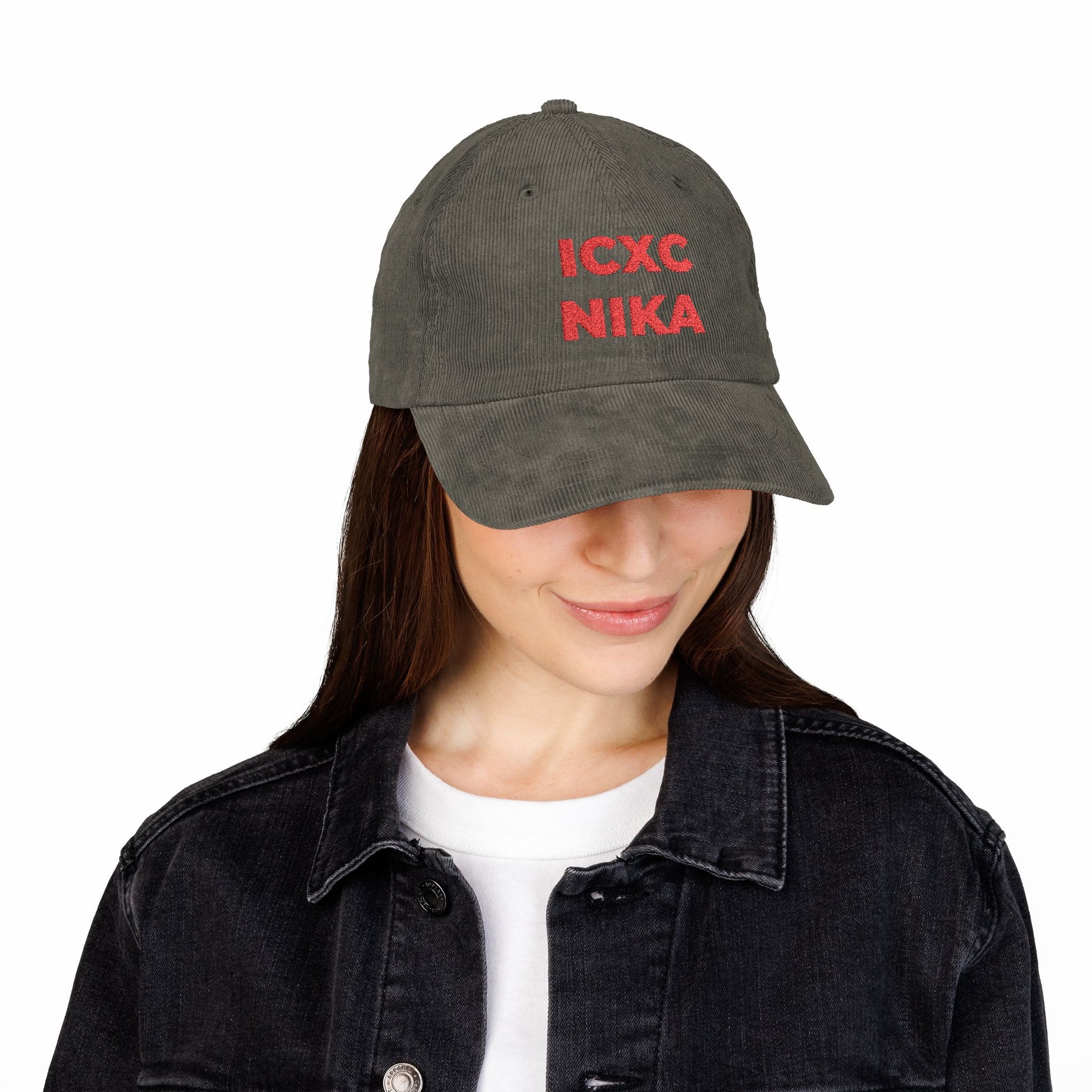A woman in a black jacket smiles as she looks down, wearing the Red ICXC NIKA Text Christogram Orthodox Christian cap—embroidered cotton corduroy featuring the symbol for "Jesus Christ Conquers.