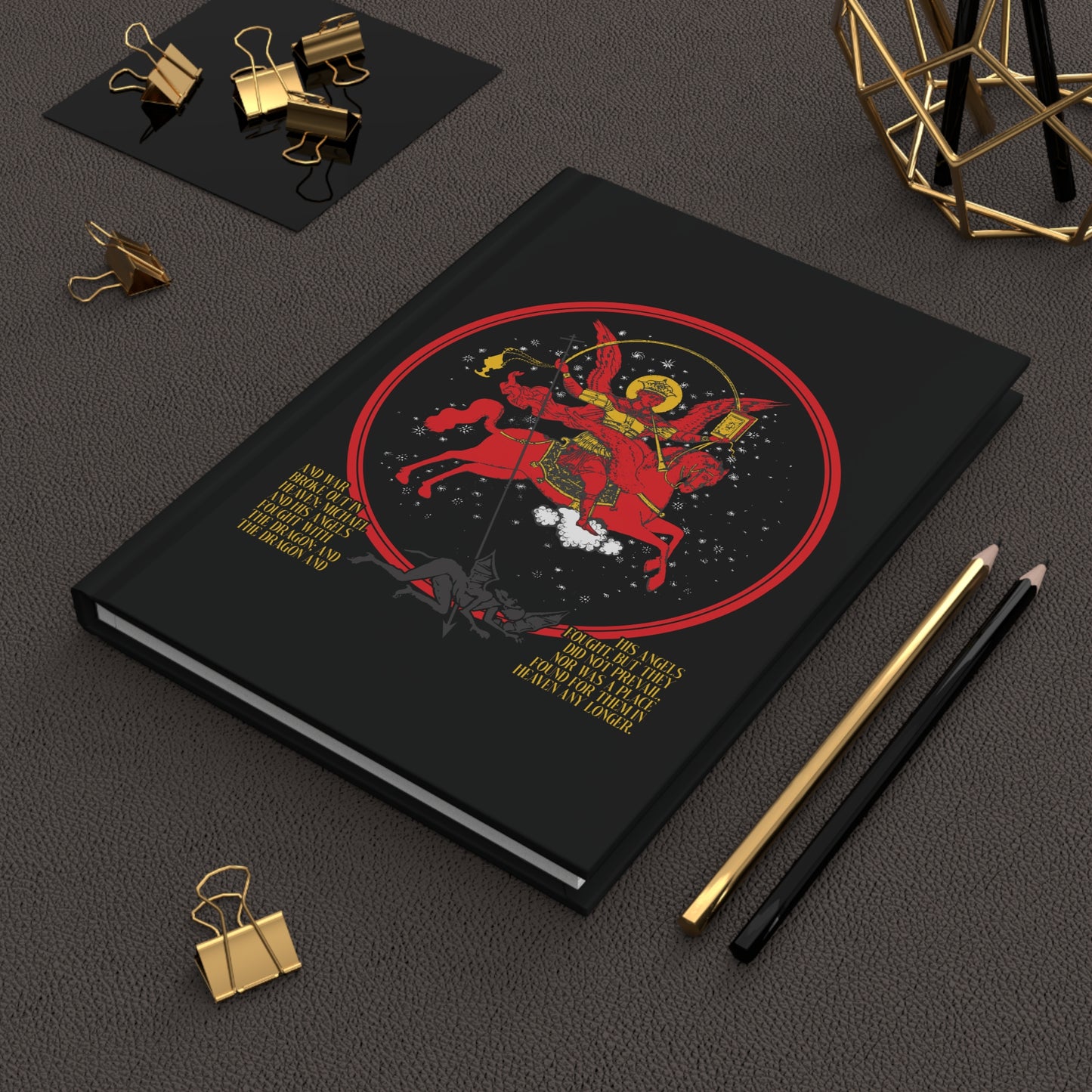 A black notebook titled "Archangel Michael of the Apocalypse Icono-Graphic | War in Heaven (Rev 12:7-9) | Satan Cast Out | Orthodox Christian Journal" lies beside pencils and binder clips.