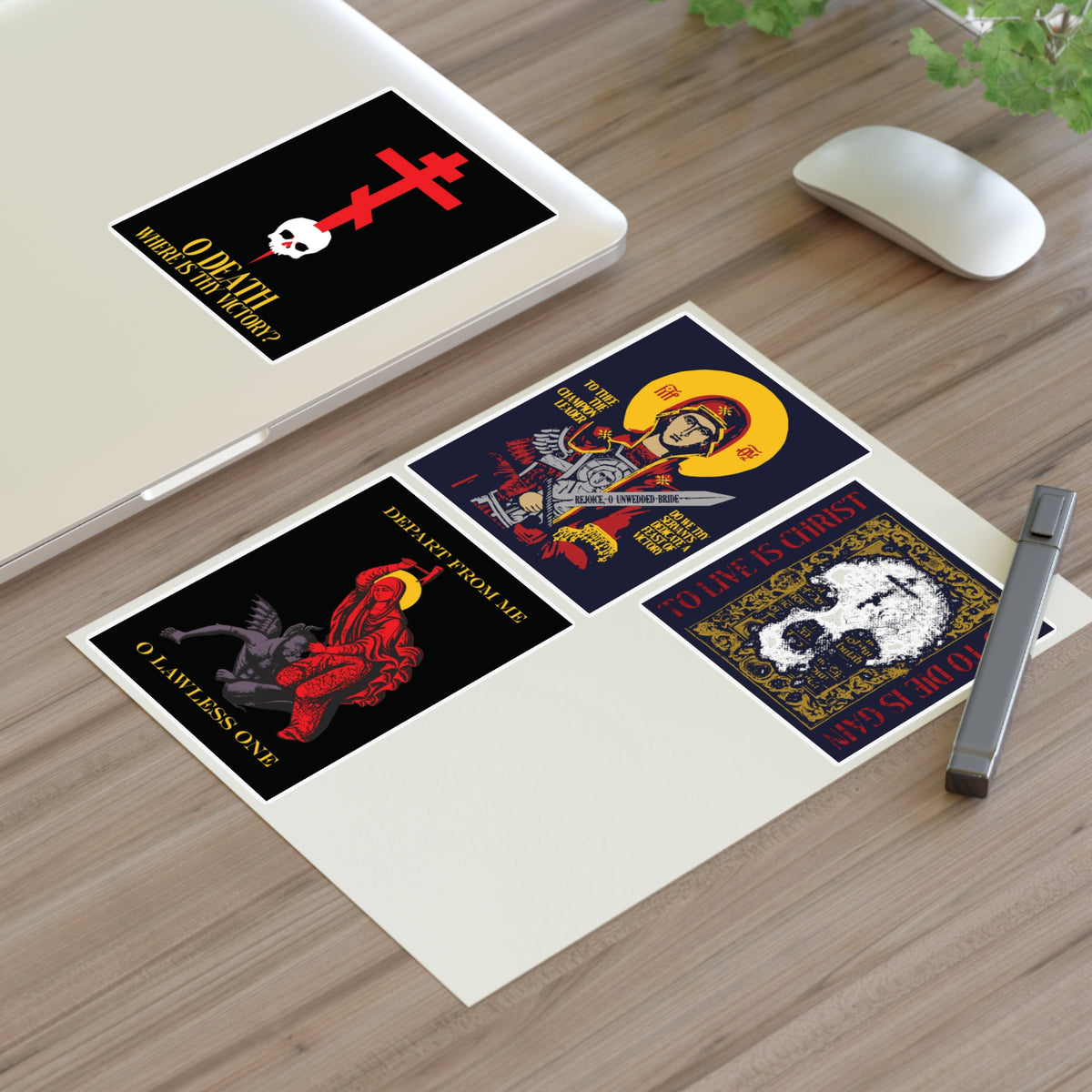 Four Orthodox stickers from the Bestseller Mix No. 1 set—including Theotokos Champion Leader, St Marina, Death Where is Thy Victory, and To Live is Christ—rest alongside a pen on a wooden desk beside a laptop and mouse, offering daily inspiration and gentle remembrance of mortality.