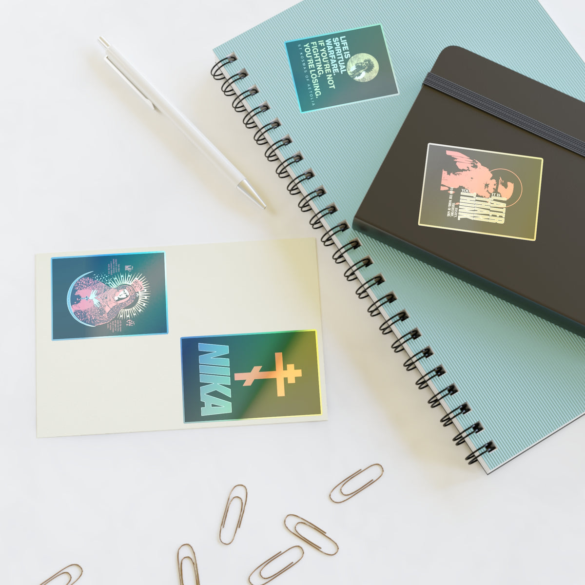 Spiral notebooks, a pen, Bestseller Orthodox Sticker Mix No. 5, and paperclips on a white surface.
