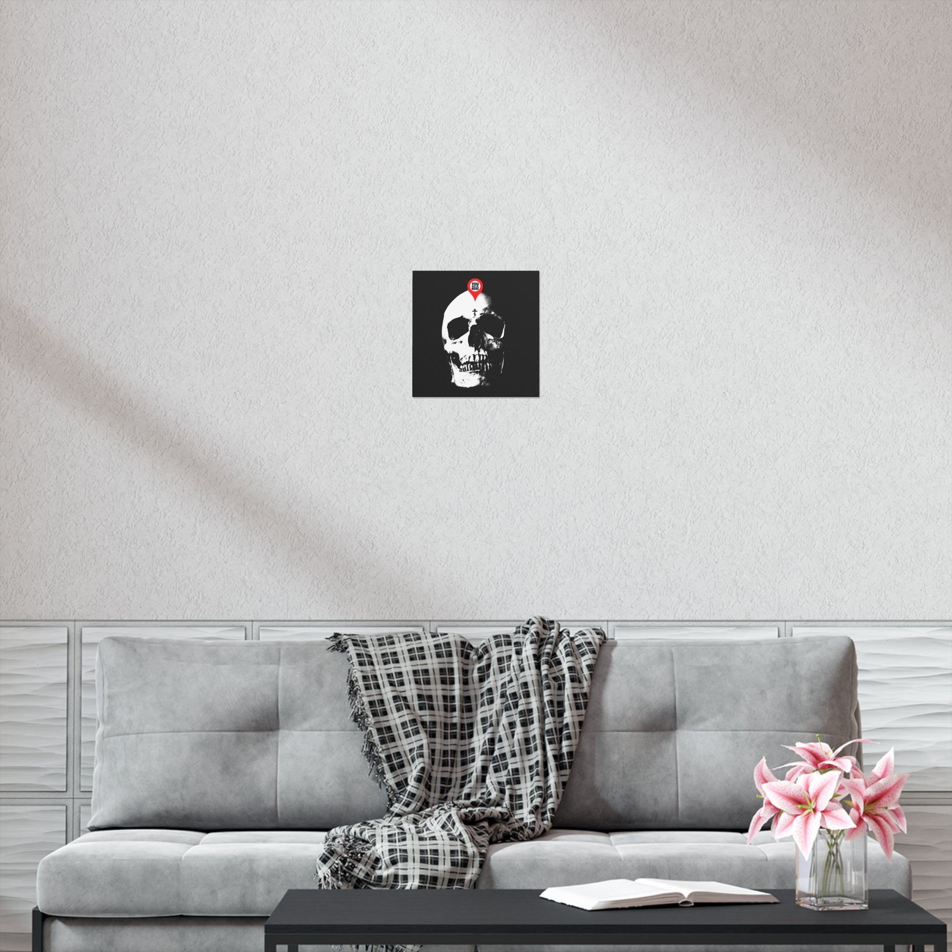 A gray sofa with a plaid throw, a coffee table with flowers and a book, and the "You Will Be Here Maps Marker on Skull Design" Orthodox Christian art poster inspire mindful reflection while elevating your decor.
