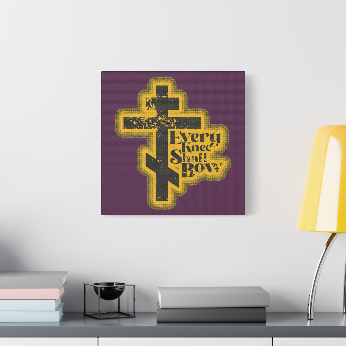 A "Every Knee Shall Bow" Christian wall art with a 70s distressed halftone comic book aesthetic, featuring Philippians 2:10-11 and Isaiah 45:23, hangs above a desk on a white wall.