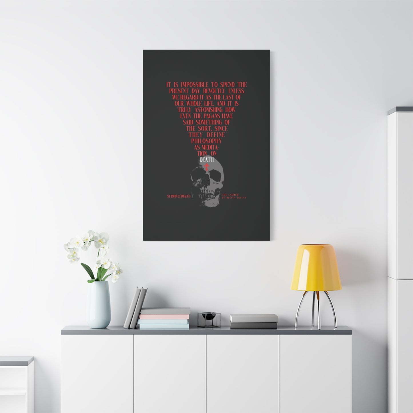 A modern room with a white sideboard, lamp, flowers, books, and "The Present Day As the Last Day of Our Life" Orthodox canvas wall art featuring a skull and red cross reflects St John Climacus’s memento mori meditation.