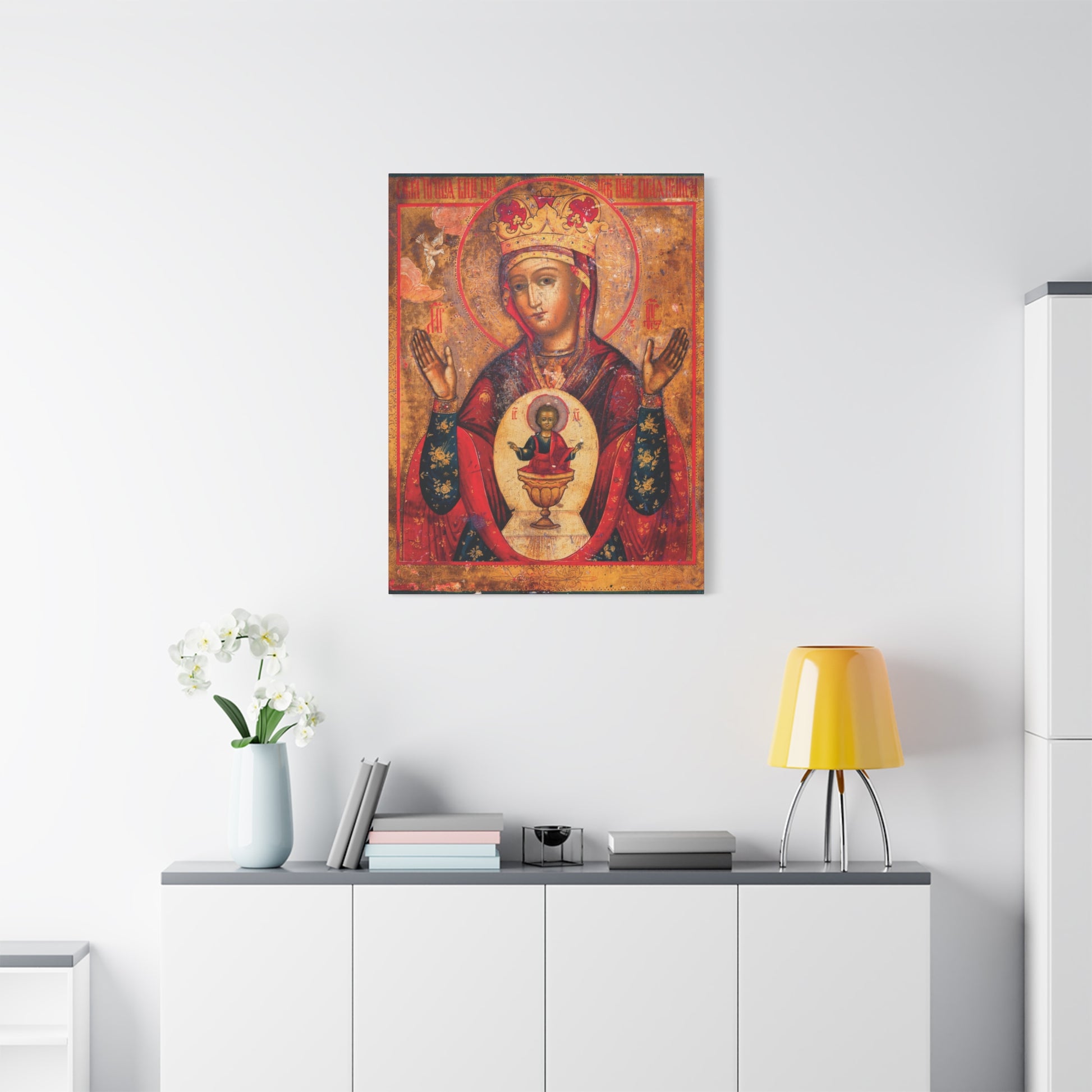 A serene vignette reminiscent of a Holy Table is created by the Mother of God of Nicaea Russian Orthodox Christian Icon | Panagia | Theotokos Mary | Religious Canvas Sacred Wall Art hanging above a white cabinet adorned with books, flowers in a vase, and a yellow lamp.