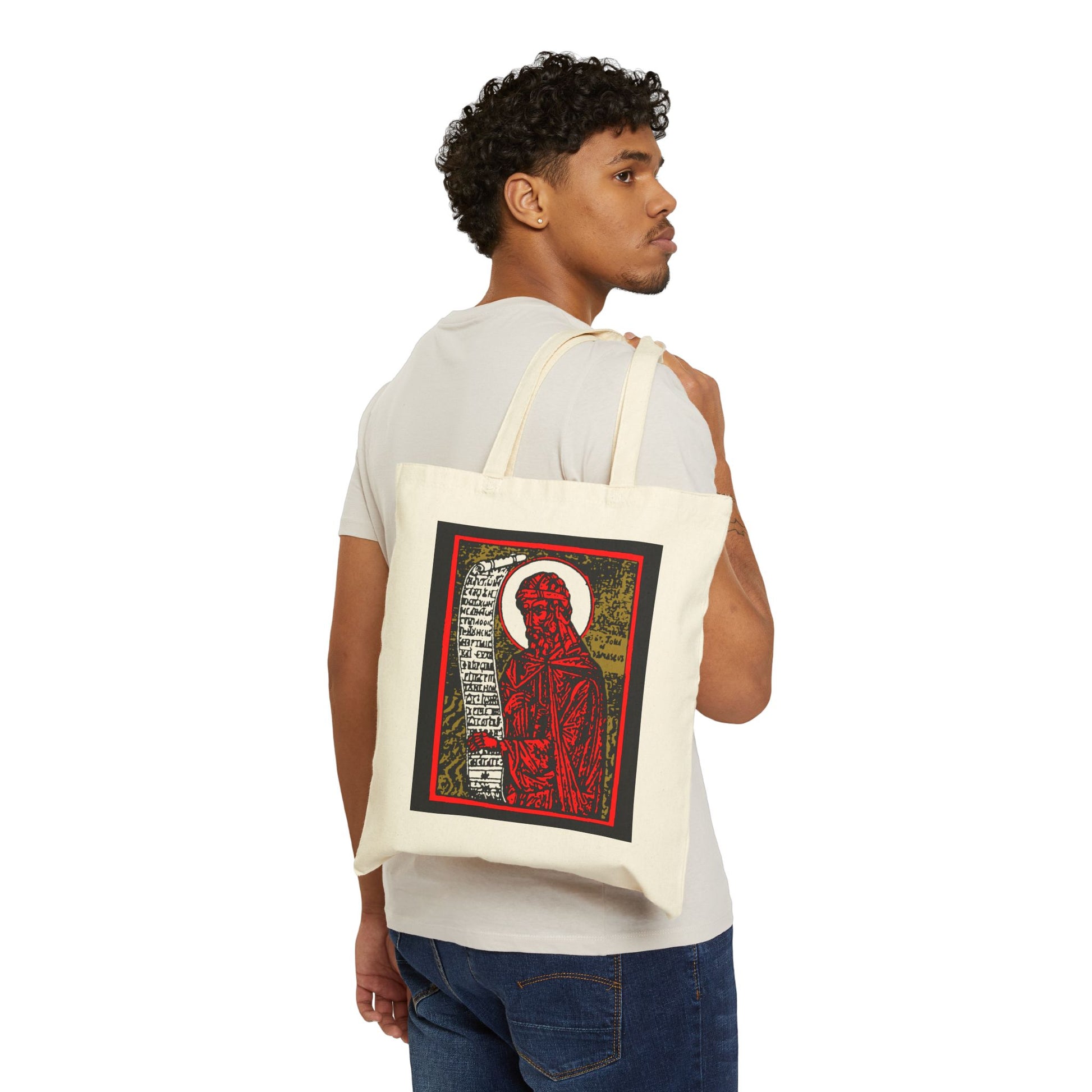 Man with curly hair holds a St. John of Damascus Icono-Graphic tote bag, featuring a red, gold & black high-contrast Orthodox Christian saint design, with text beside the haloed figure. Cotton construction.