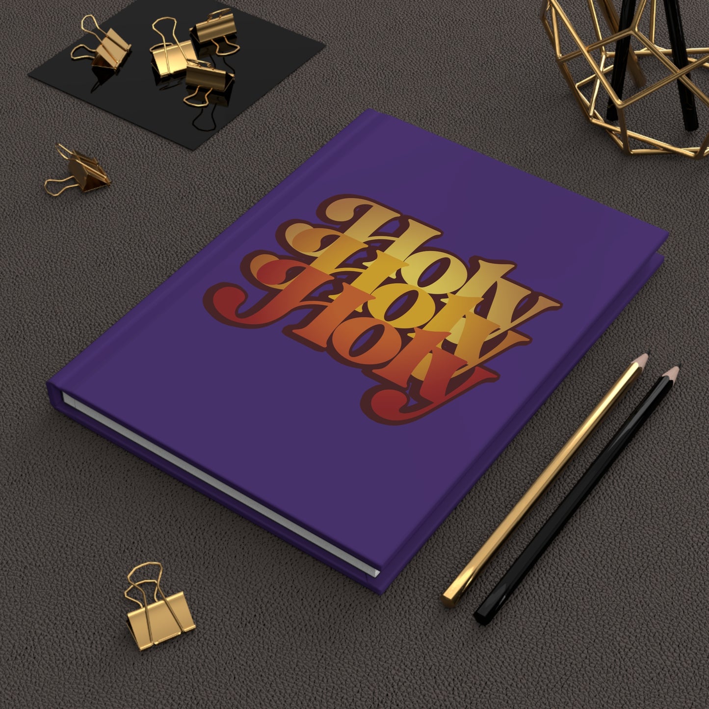 A purple "Holy Holy Holy Trisagion Liturgical Prayer" hardcover journal with a 70s style and Isaiah 6 design rests next to pencils, clips, and geometric decor.