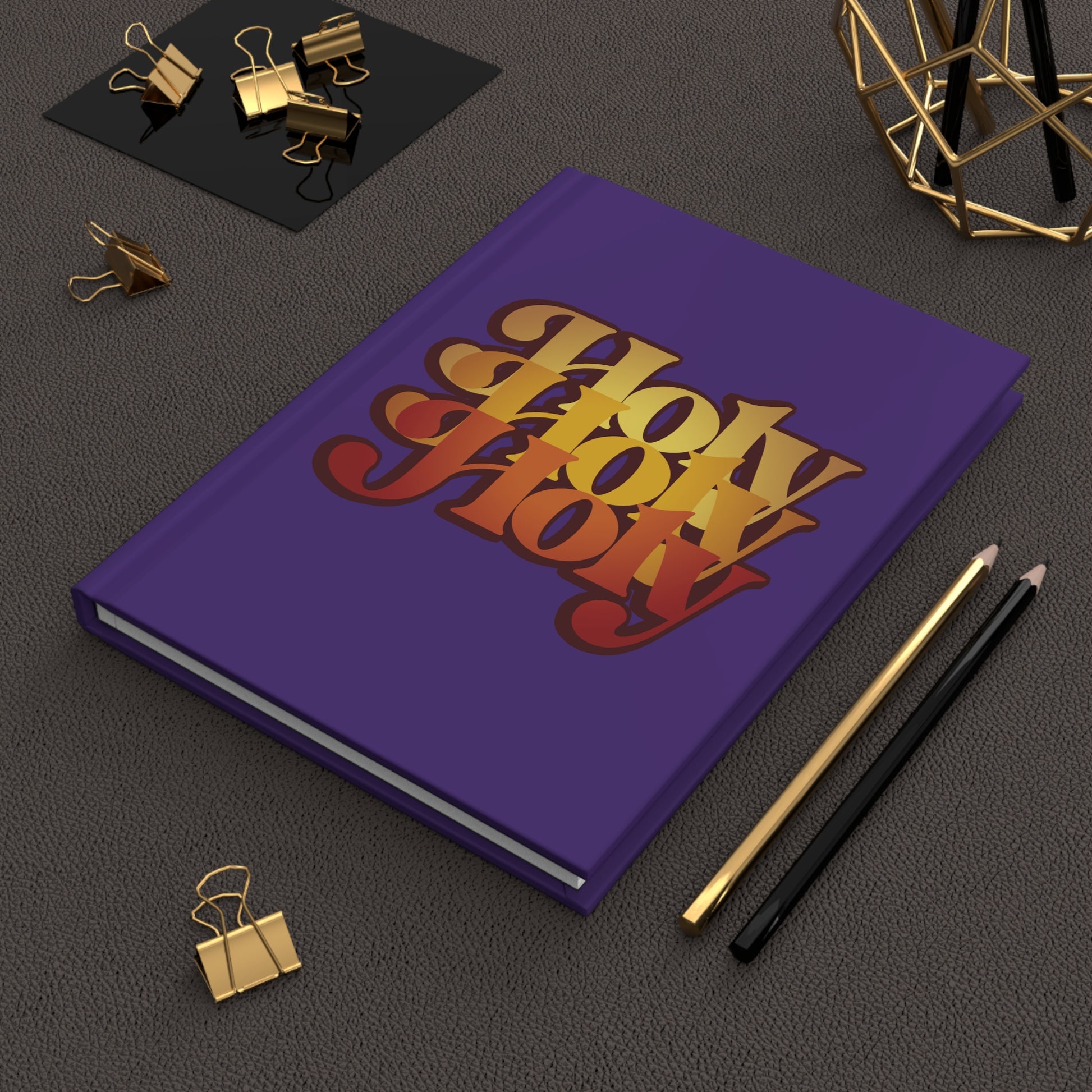 A purple "Holy Holy Holy Trisagion Liturgical Prayer" hardcover journal with a 70s style and Isaiah 6 design rests next to pencils, clips, and geometric decor.