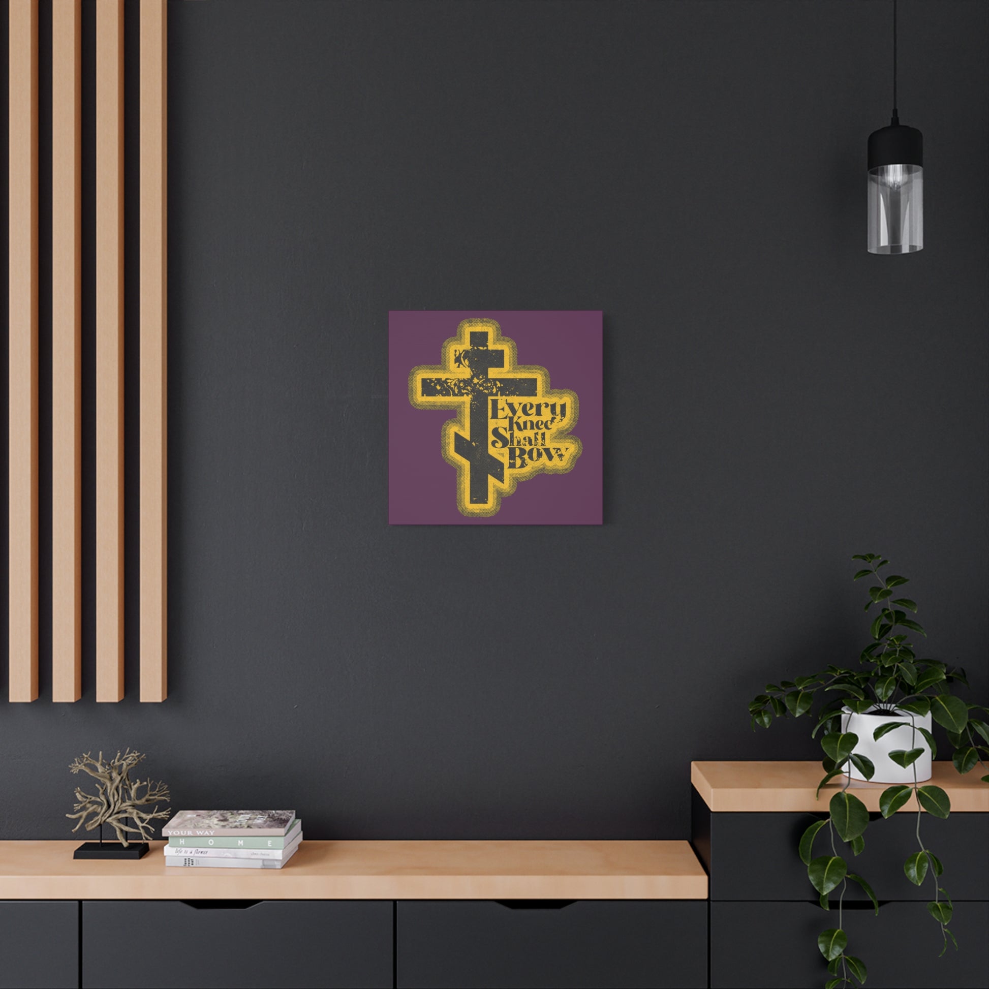 A modern room with dark walls and wooden accents features "Every Knee Shall Bow | 70s Distressed Halftone Comic Book Aesthetic | Philippians 2:10-11 | Isaiah 45:23" Christian Wall Art above a black cabinet with plants.