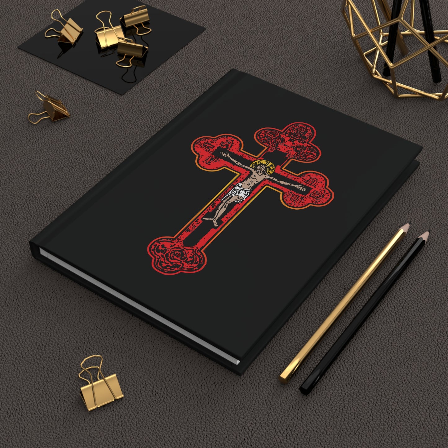 The "Red Orthodox Christian Crucifix | Byzantine Golgotha Cross" hardcover journal, inspired by "I Am Crucified With Christ," rests beside gold binder clips and two pencils on a brown surface.