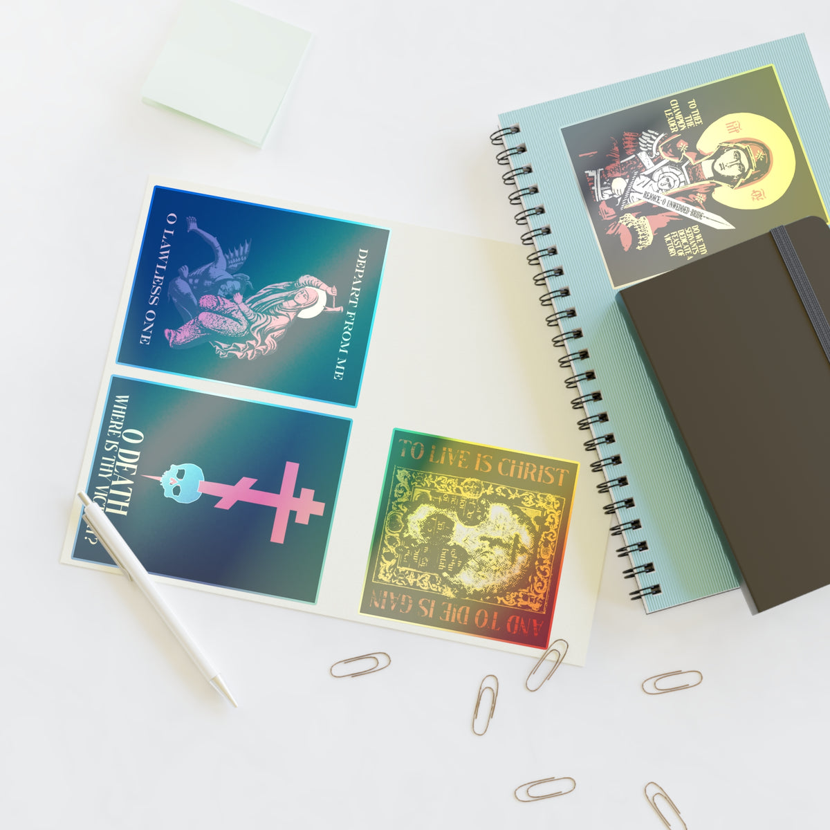Open notebook with colorful religious-themed cards, Bestseller Mix No. 1 Four Orthodox Stickers (featuring Theotokos Champion Leader, St Marina, Death Where is Thy Victory, and To Live is Christ), pen, sticky notes, and paper clips on a white desk.