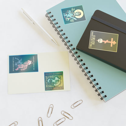 On a white desk are two notebooks, a pen, a postcard, and paper clips—all decorated with Orthodox-themed stickers from the Bestseller Mix No. 1 set, featuring designs such as Theotokos Champion Leader, St Marina, Death Where is Thy Victory, and To Live is Christ.