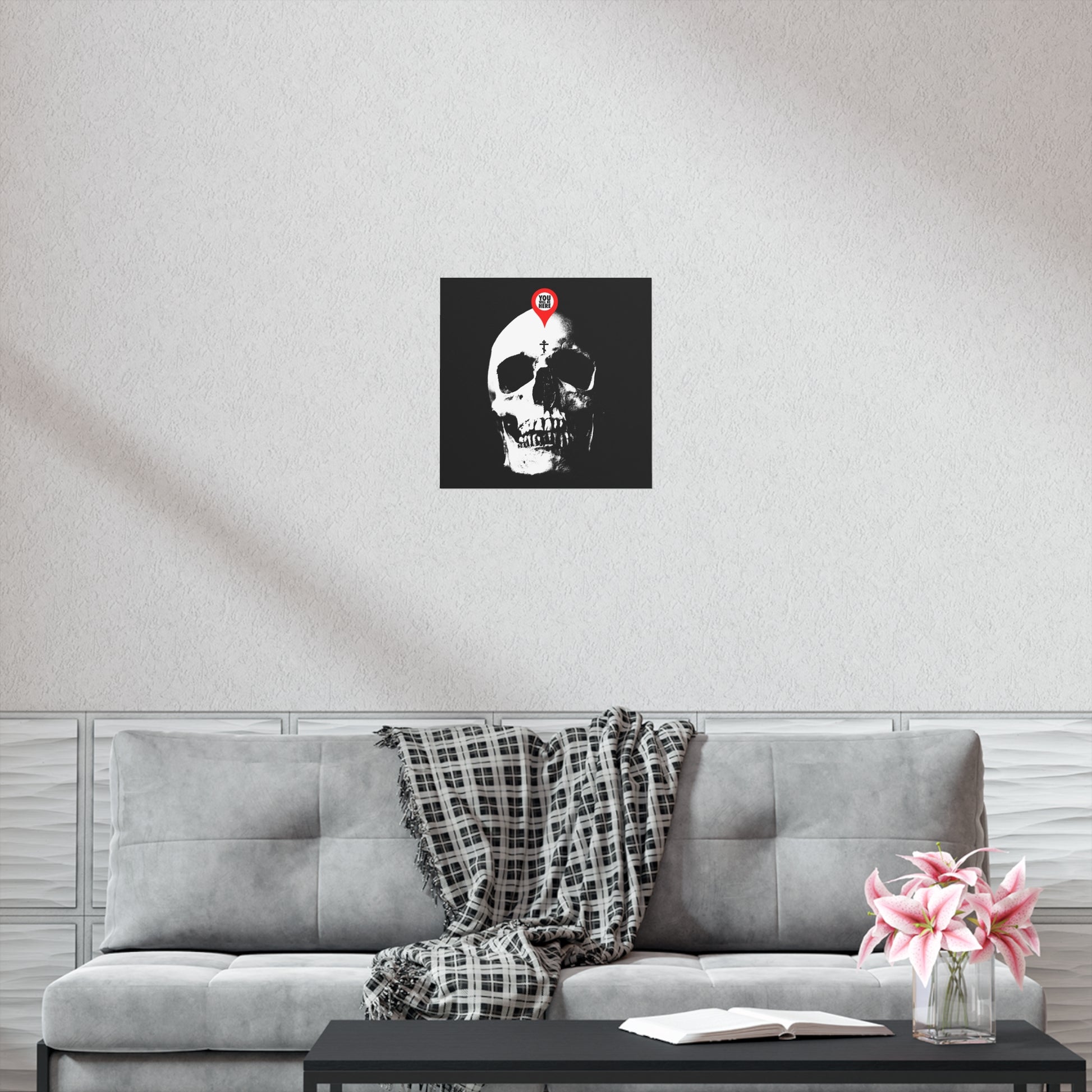 A gray couch with a plaid blanket, a pink flower vase, and the "You Will Be Here Maps Marker on Skull Design" Orthodox Christian art poster above—a memento mori echoing St Ignatius Brianchaninov’s teachings.