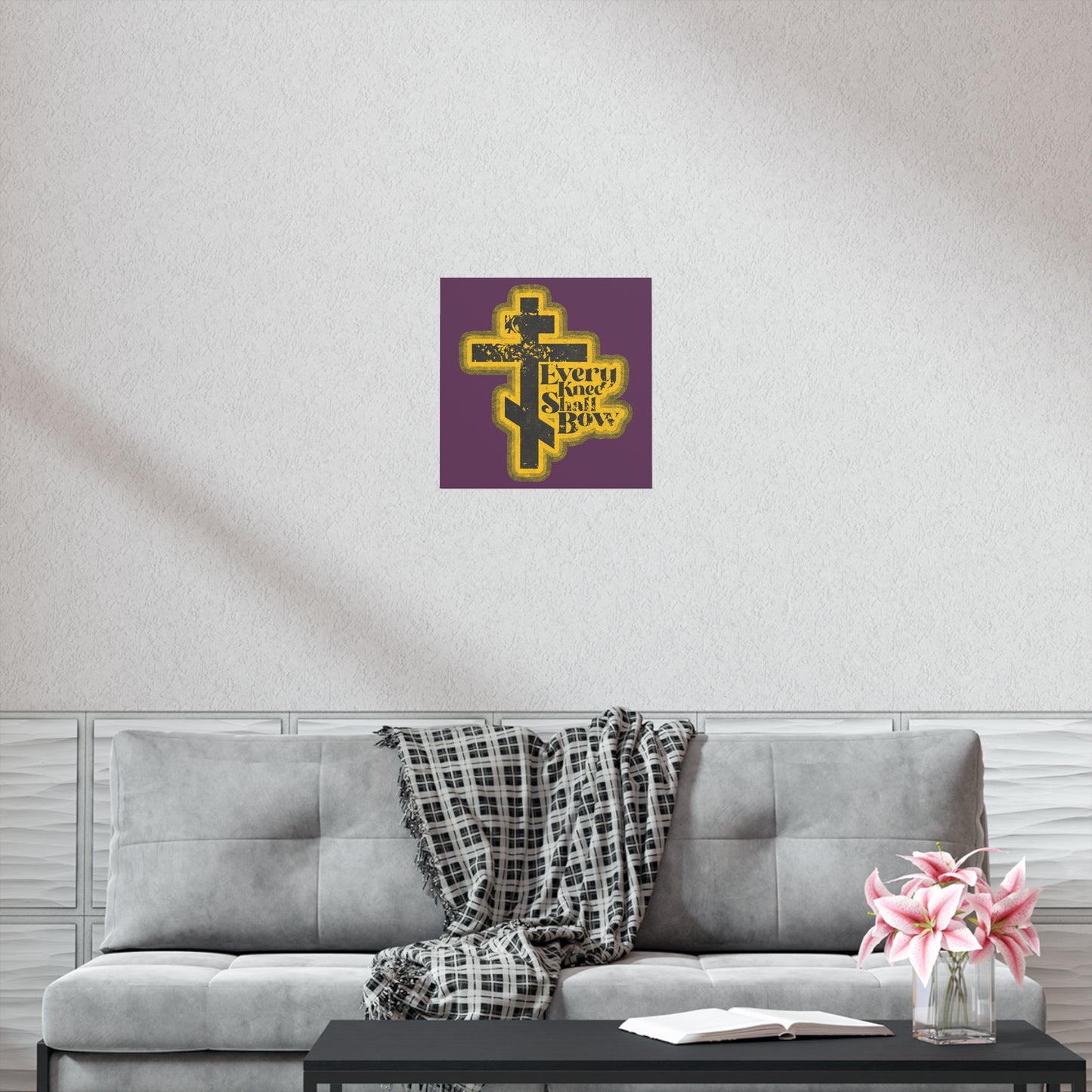 A modern living room features the "Every Knee Shall Bow" Orthodox Christian art poster with a 70s distressed halftone comic book style, inspired by Philippians 2:10-11 and Isaiah 45:23, above a gray sofa with flowers and a checkered throw.
