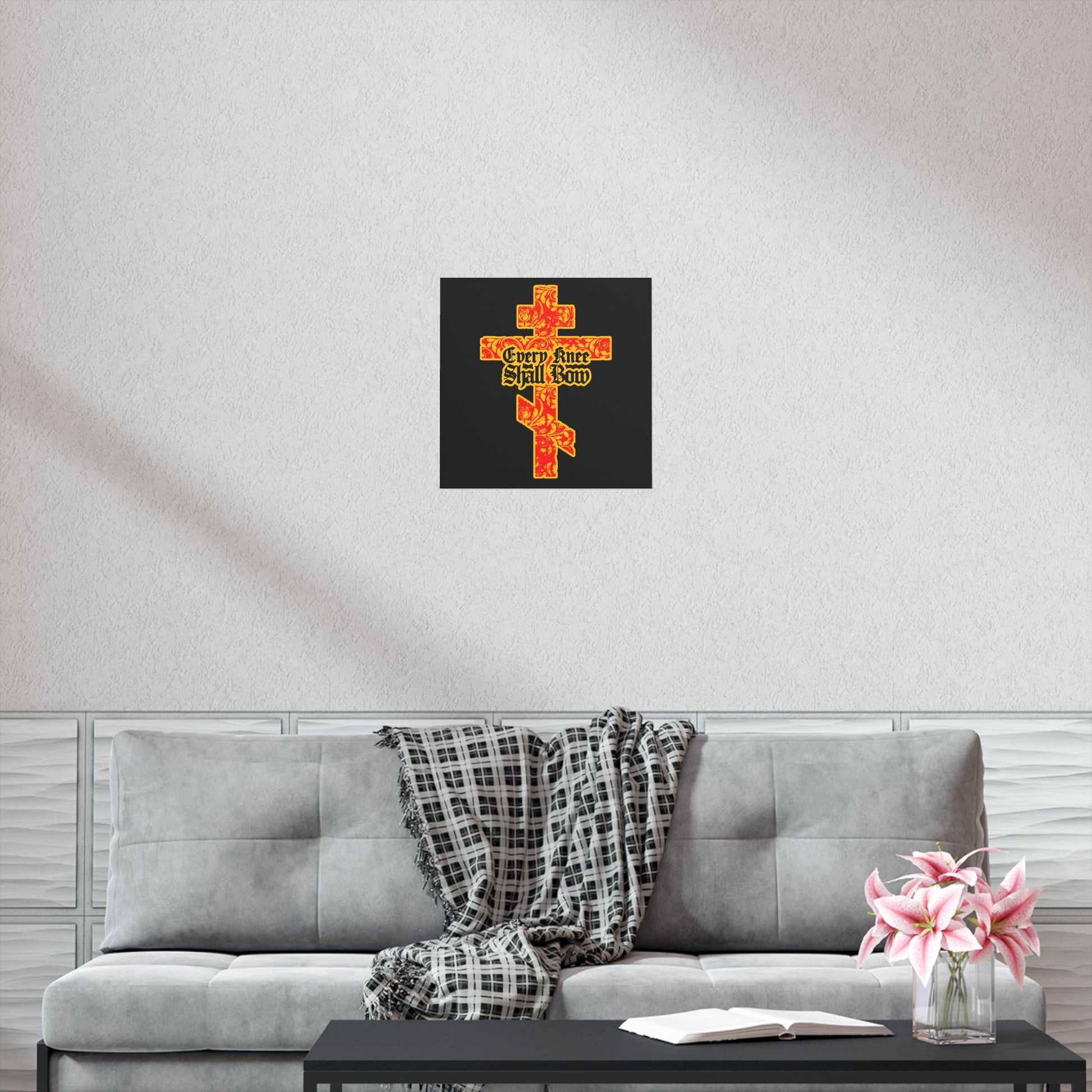 A grey sofa with a plaid throw, pink flowers, and a book sits beneath the "Every Knee Shall Bow" gold and red floral Orthodox Cross art poster, creating a peaceful space inspired by faith’s strength and righteousness.
