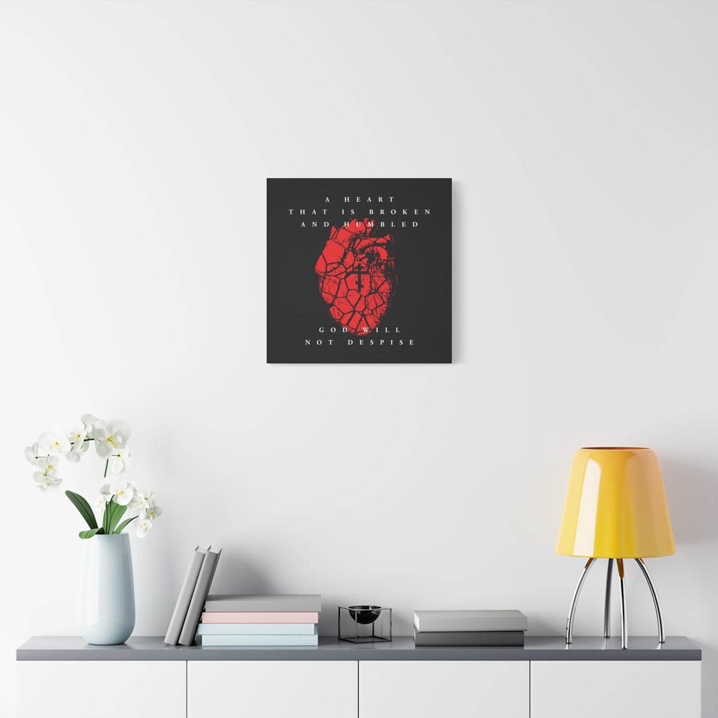 A Christian canvas art, "A Heart That is Broken and Humbled God Will Not Despise," featuring a cracked heart with an Orthodox cross, hangs above a white cabinet adorned with a lamp, vase, books, and flowers.