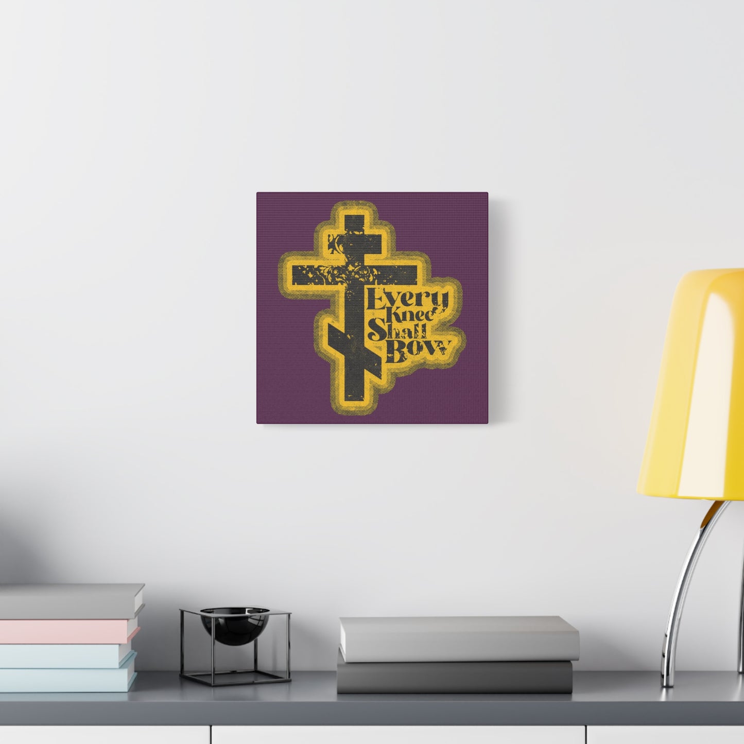 A purple wall displays the "Every Knee Shall Bow | 70s Distressed Halftone Comic Book Aesthetic | Philippians 2:10-11 | Isaiah 45:23 | Christian Wall Art" above a shelf decorated with books and accents.