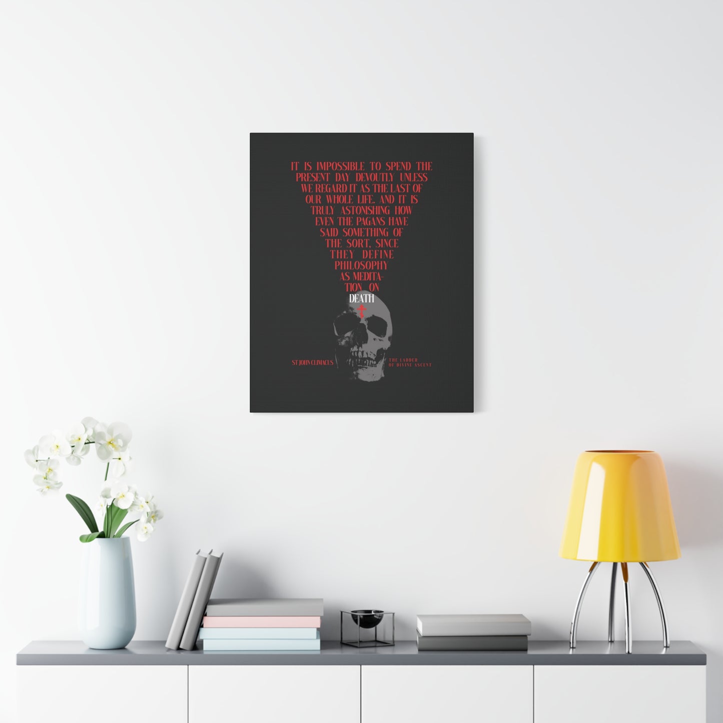 A modern room featuring "The Present Day As the Last Day of Our Life" Orthodox canvas wall art with a skull and red cross, displayed above a white console with flowers, books, and a yellow lamp.