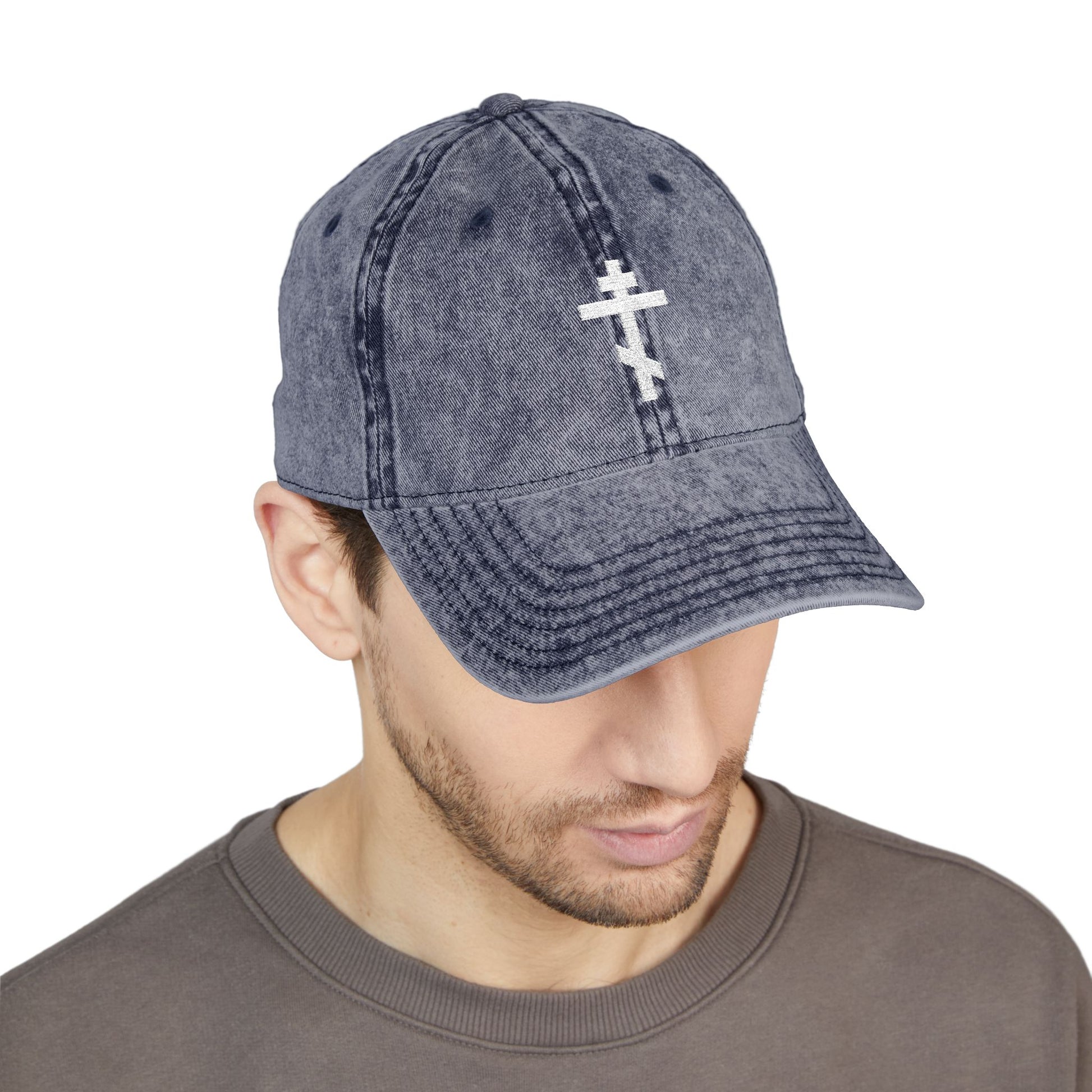 Man wearing a Simple White Orthodox TriBar Cross cap—Minimalist Christian distressed gray denim hat with white embroidered cross and "Crucify My Ego to the Precious and Life Giving Cross" message—looking downward.