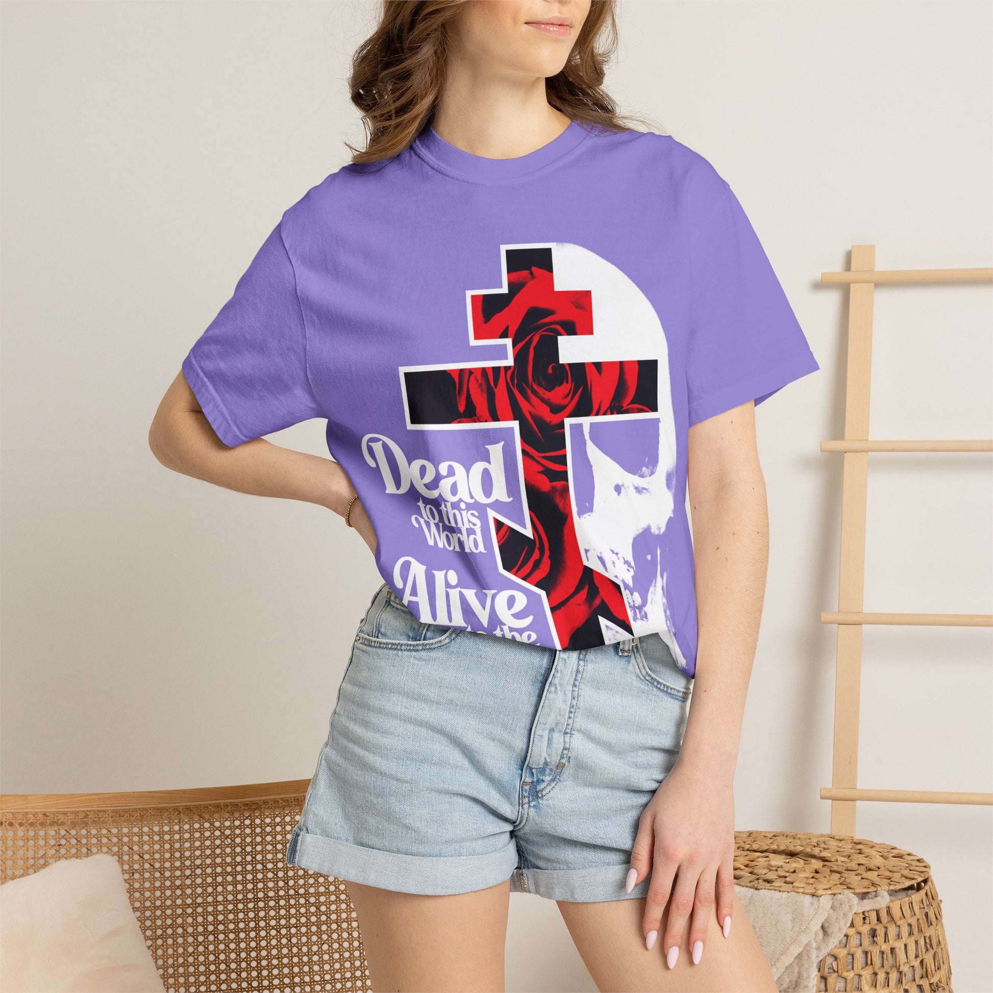 A woman wearing the "Dead to this World" Christian Goth Flower Cross & Skull Heavyweight Cotton Unisex T-Shirt with 70s typography and denim shorts stands indoors.