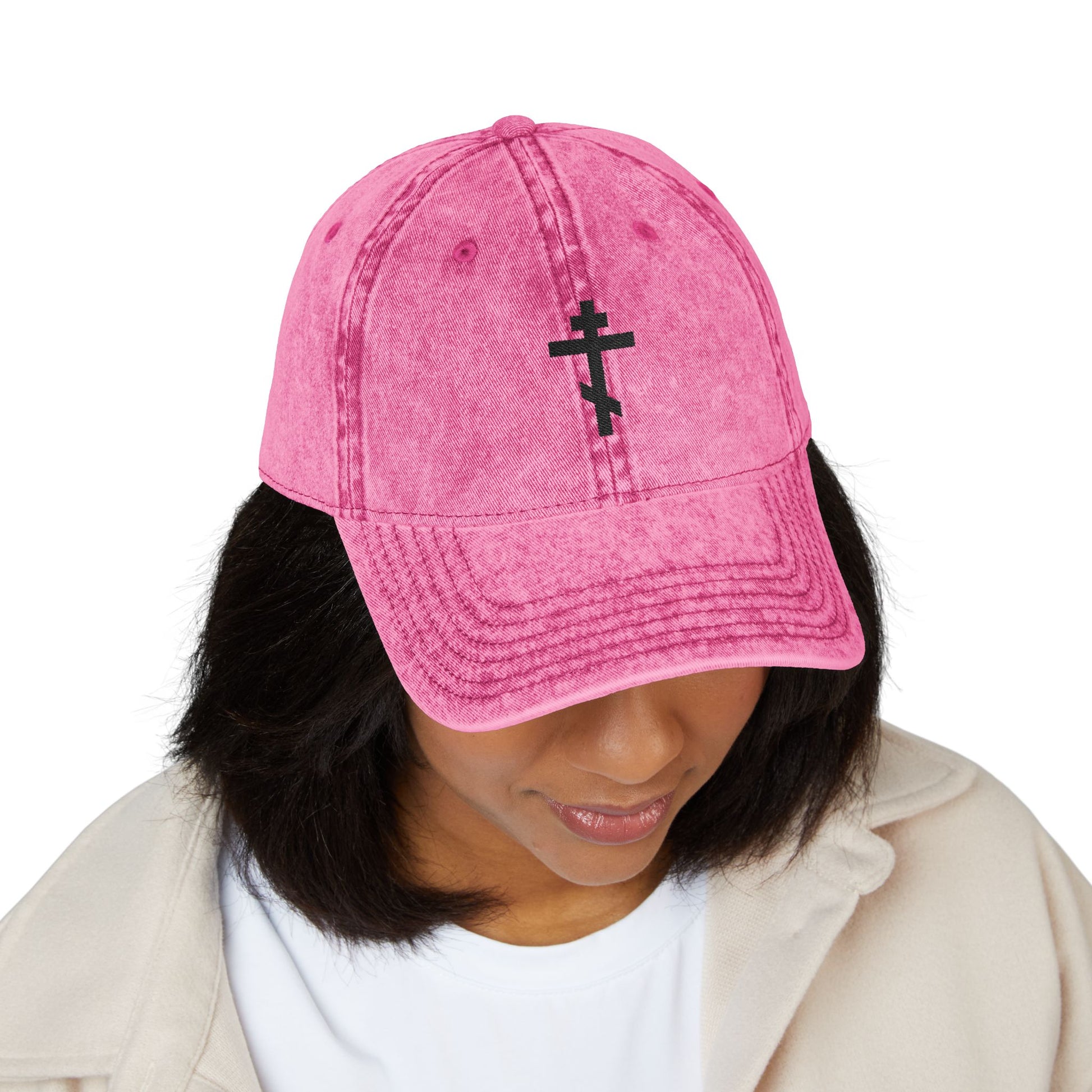 Woman wearing the Simple Black Orthodox TriBar Cross Cap—pink, minimalist, with a black distressed embroidered Crucify My Ego to the Precious and Life Giving Cross symbol—looking down.