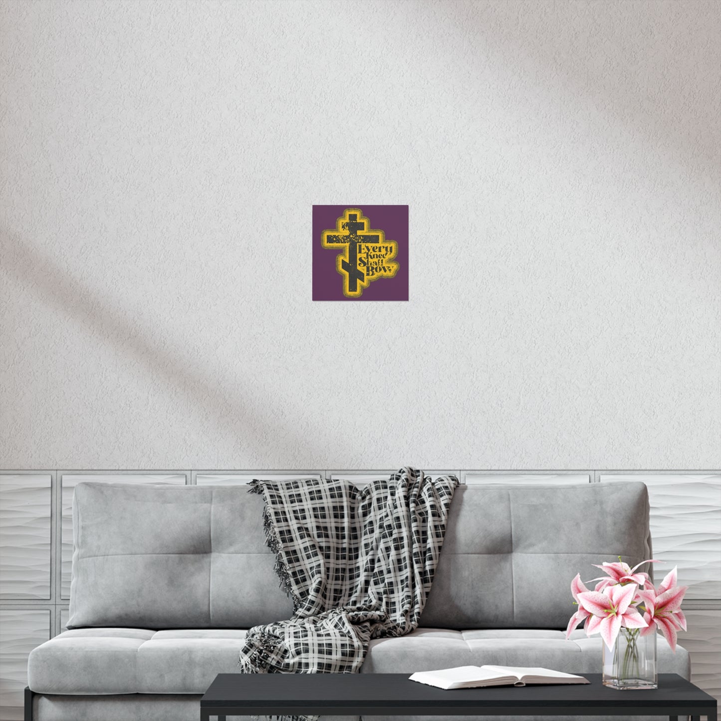 A cozy living room with a grey couch, plaid throw, coffee table, flowers, and the "Every Knee Shall Bow" 70s distressed comic-style Orthodox Christian art poster (Philippians 2:10-11) displayed above the sofa.