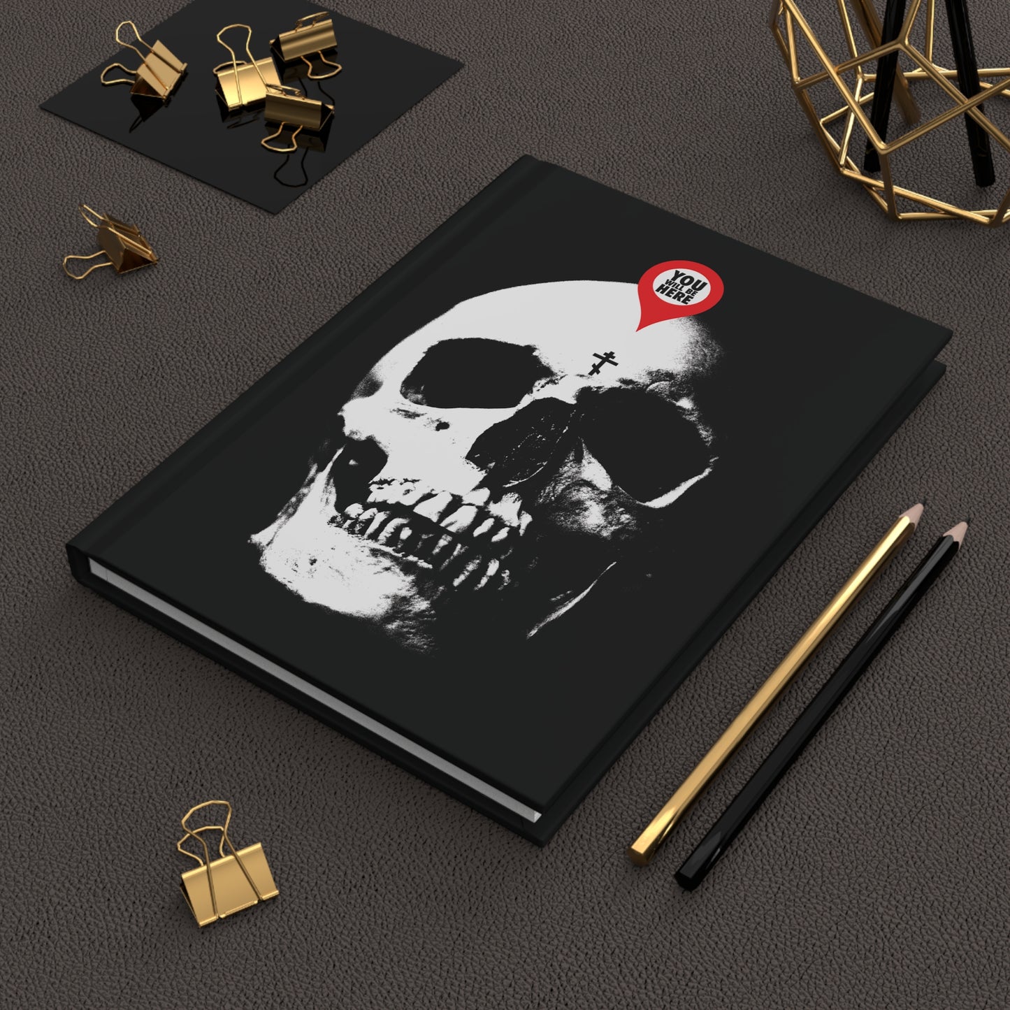 The "You Will Be Here Maps Marker on Skull Design" Orthodox Blank Journal, paired with two pencils and gold binder clips on a dark surface, inspires contemplation of mortality and the message: Remember You Will Die (Memento Mori).
