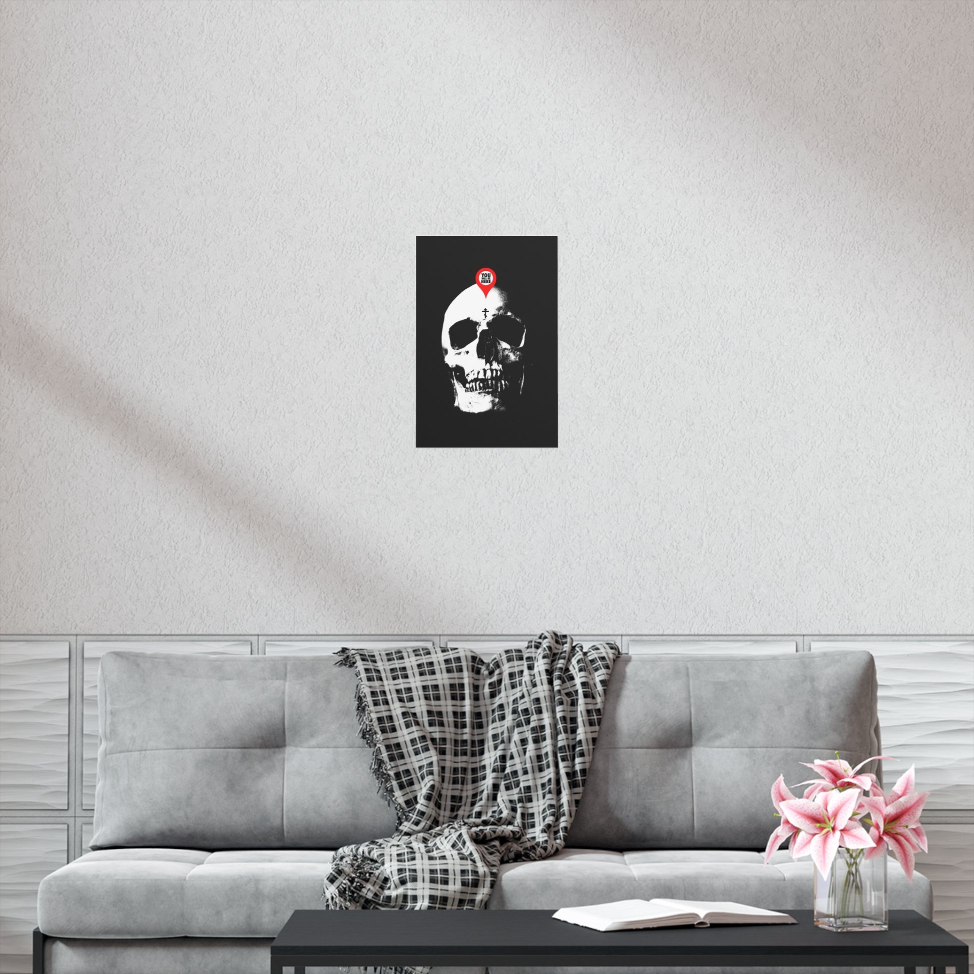 A "You Will Be Here Maps Marker on Skull" Orthodox Christian art poster, featuring the Memento Mori message, hangs above a gray sofa with a plaid throw, flowers, and an open book—echoing St. Ignatius Brianchaninov’s reflection on spiritual death.
